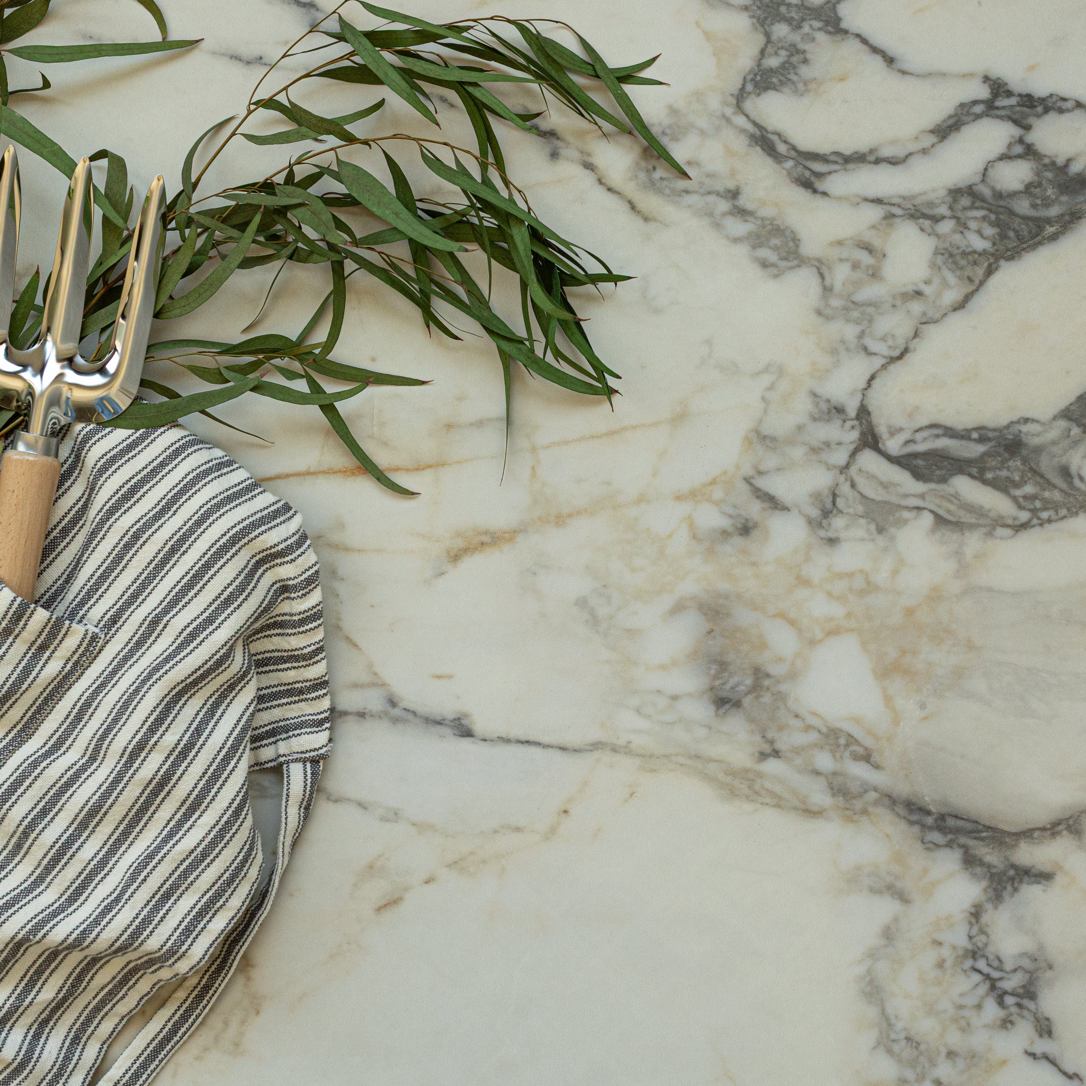 Light marble surface with gray and beige veining, garden fork with wooden handle, striped cloth, and green leafy branch