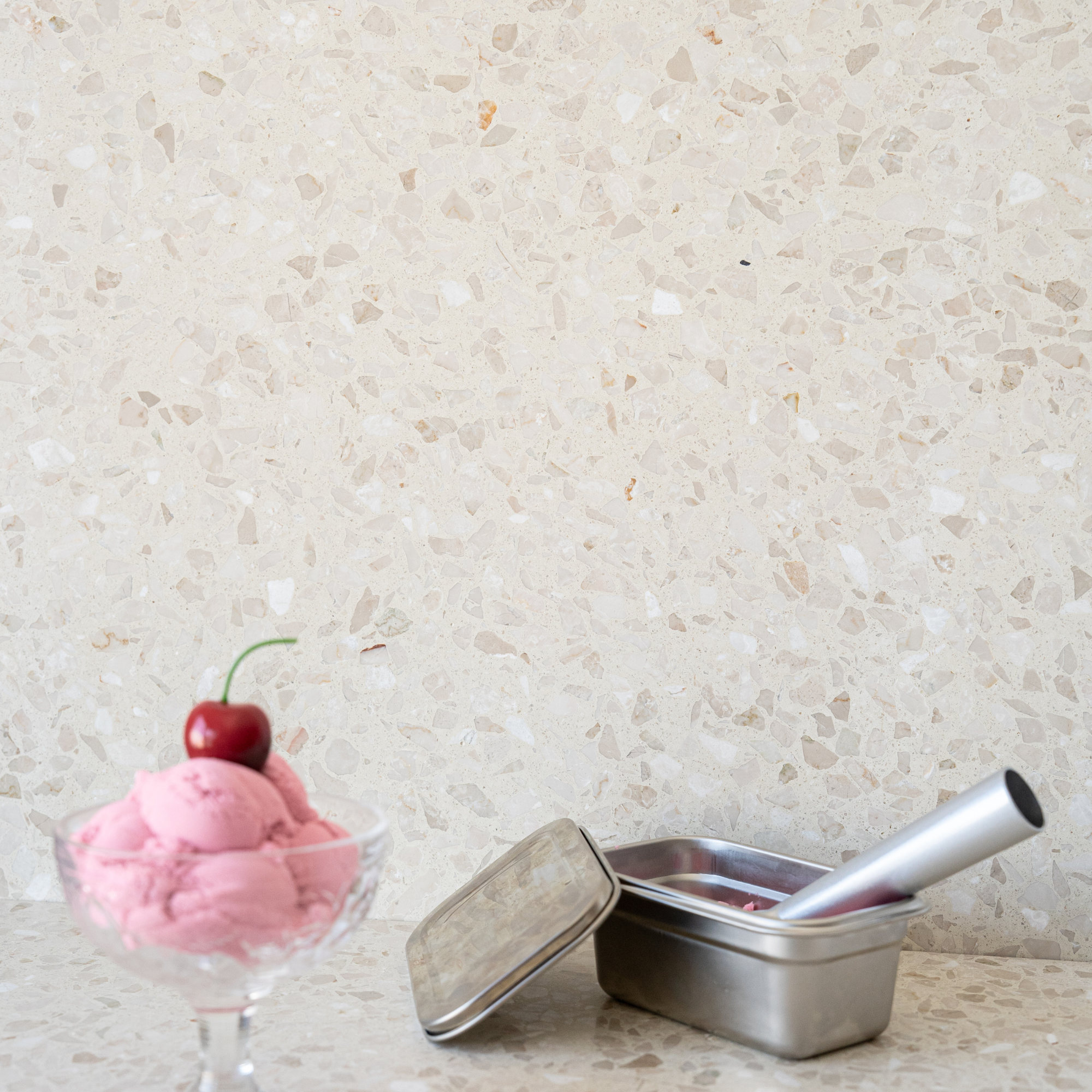 Cream terrazzo surface with pink ice cream topped by a cherry beside a metal pan and scoop
