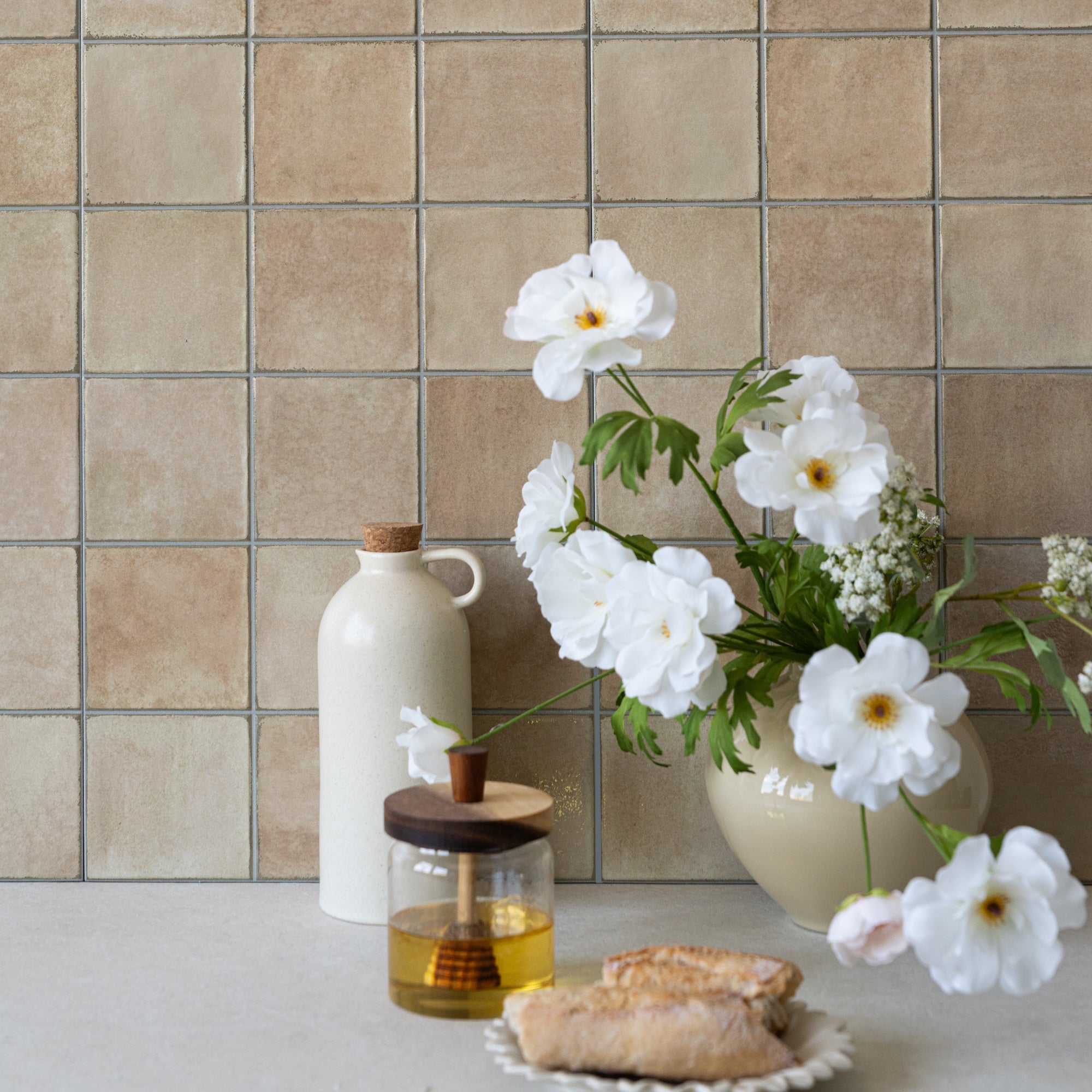 Beige ceramic tile wall with white flowers in a vase, a ceramic bottle, honey jar, and bread on countertop