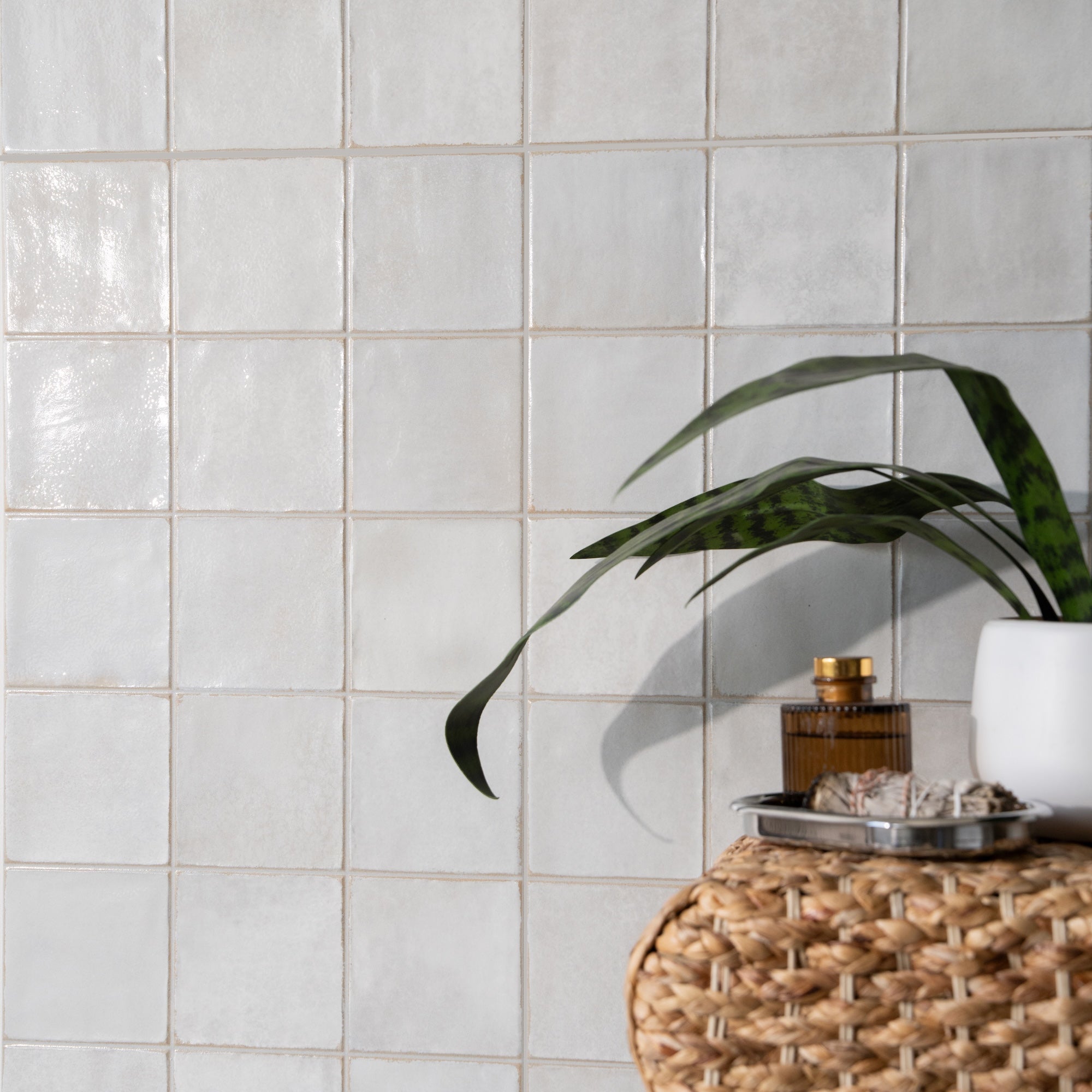 Glossy white ceramic tiles on wall with woven basket, green plant, and amber glass bottle on tray nearby