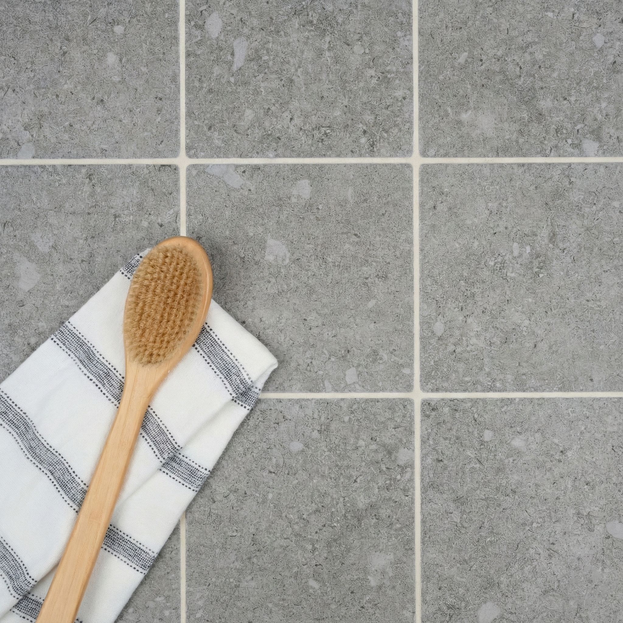Overhead view of square ash gray ceramic tiles with subtle stone texture alongside a wooden bath brush and striped cloth