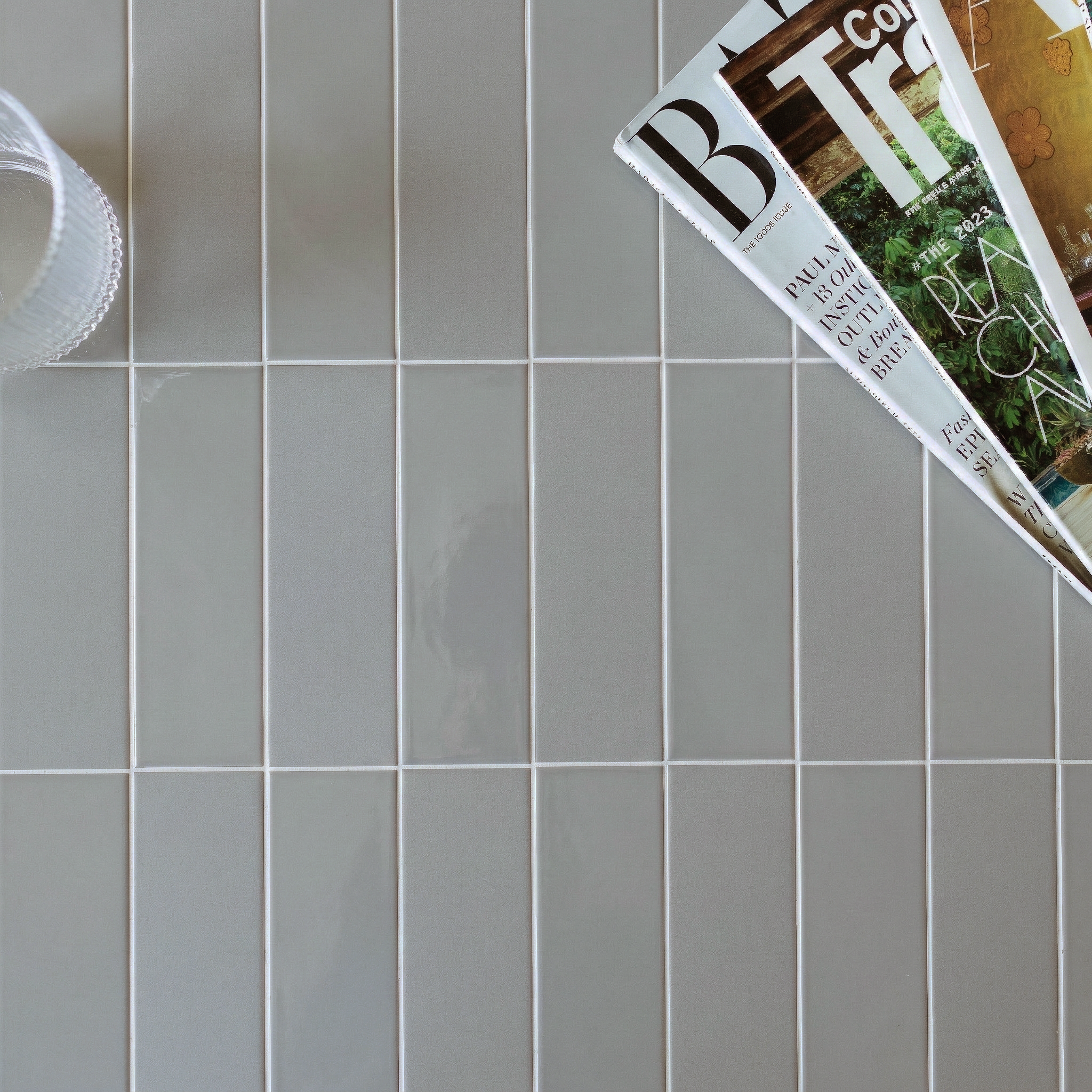 Glossy light gray rectangular ceramic tiles with white grout next to overlapping magazines and a clear glass bowl