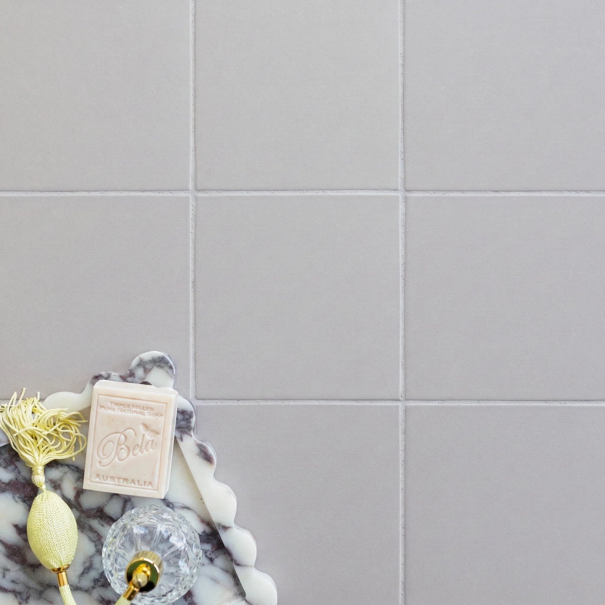 Soft matte pale ash porcelain tiles with subtle grout lines and a marble tray holding vintage perfume and soap