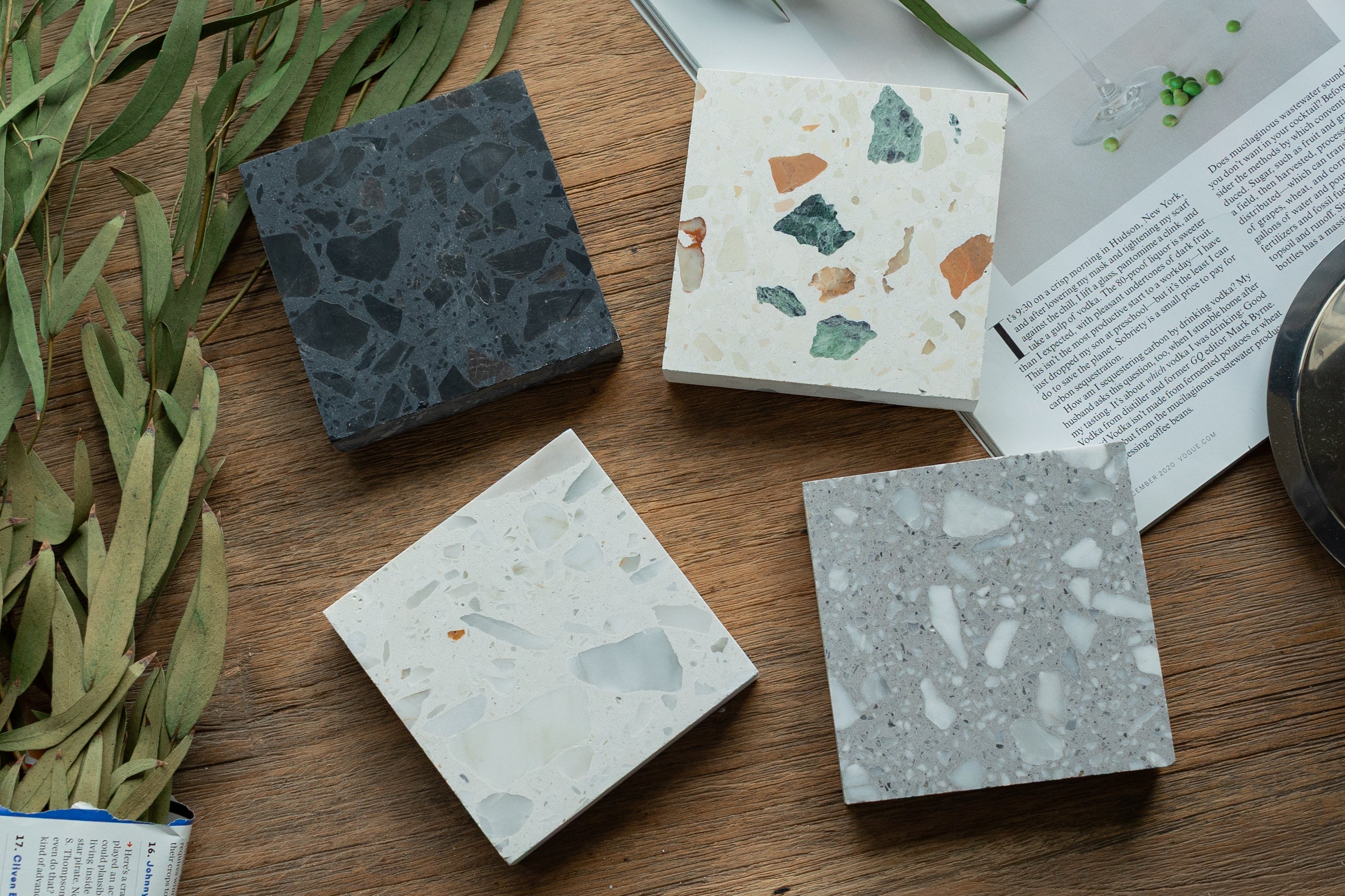 Four terrazzo square tile samples in black, white, beige, and gray on a wooden surface with papers and leaves nearby