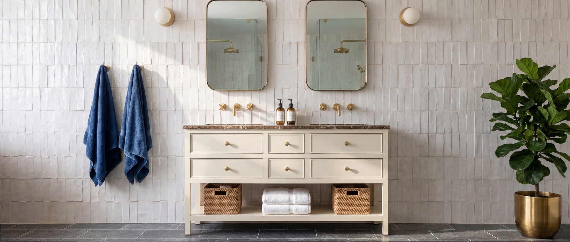 Bright bathroom with textured off-white tile wall, double oval mirrors, beige vanity, brass fixtures, and a green potted plant