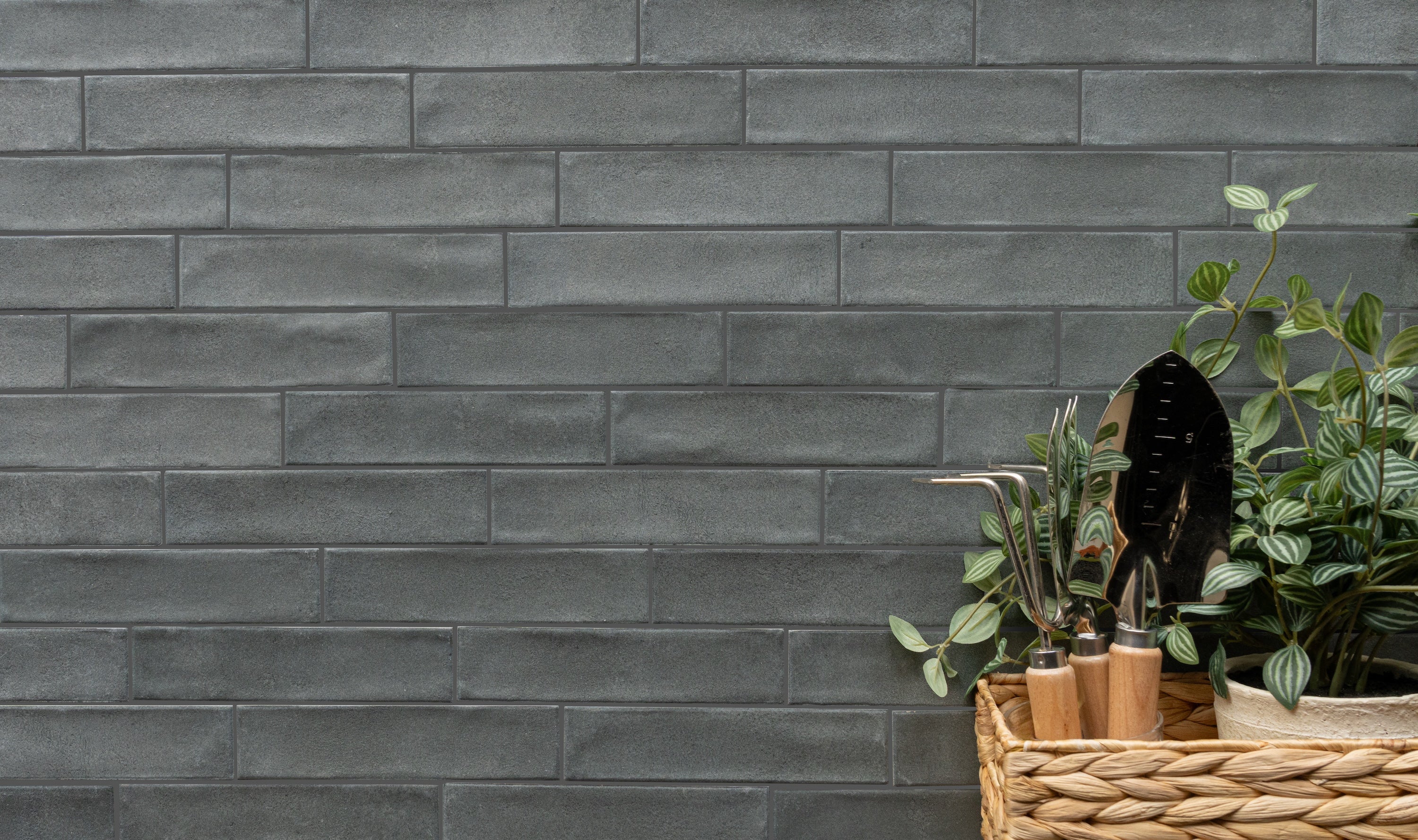 Gray rectangular ceramic tile wall with a wicker basket of gardening tools and green plants on the side