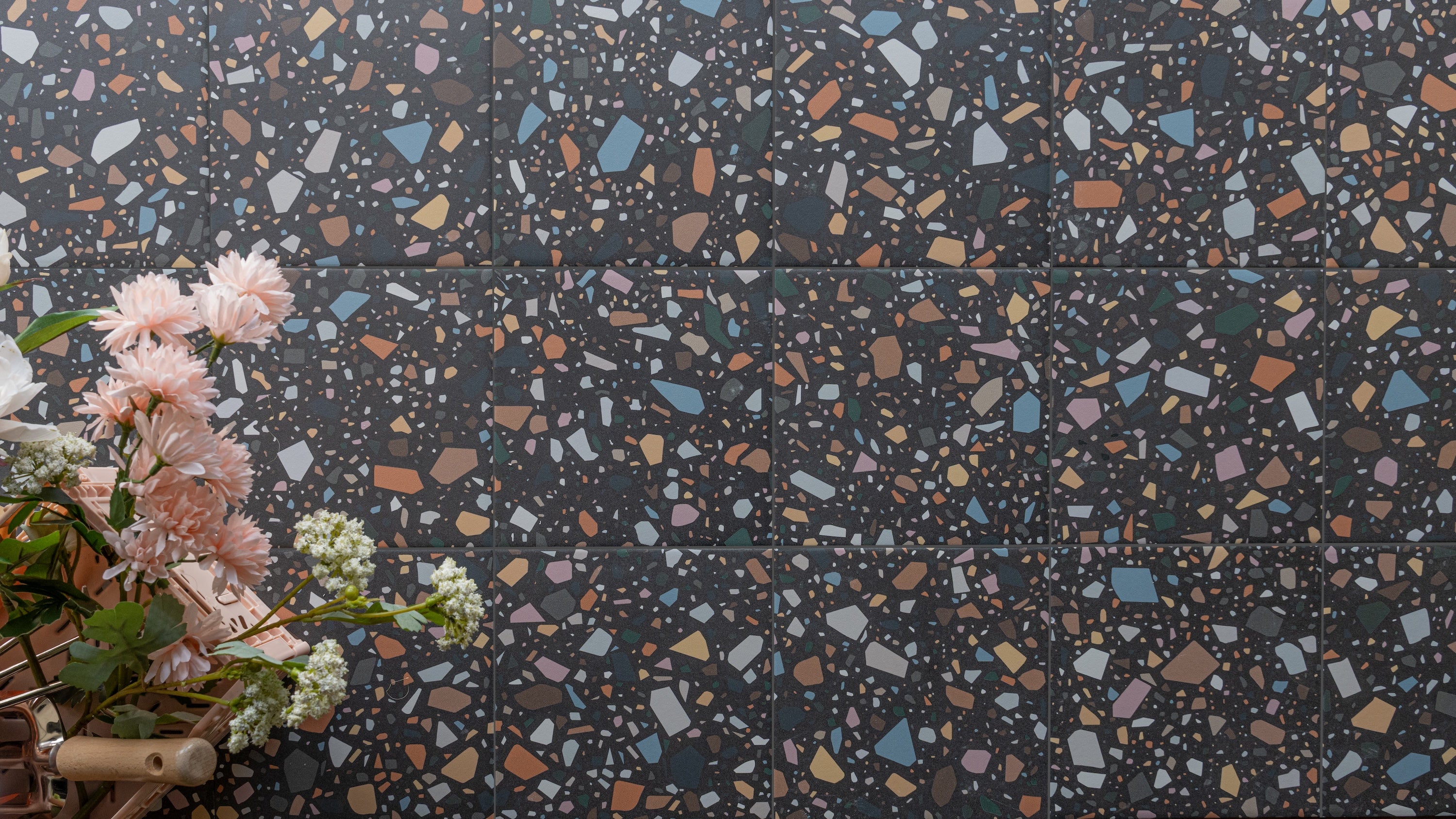 Dark terrazzo tiles with multicolored stone chips in blue, orange, and white, accompanied by soft pink flowers on the side
