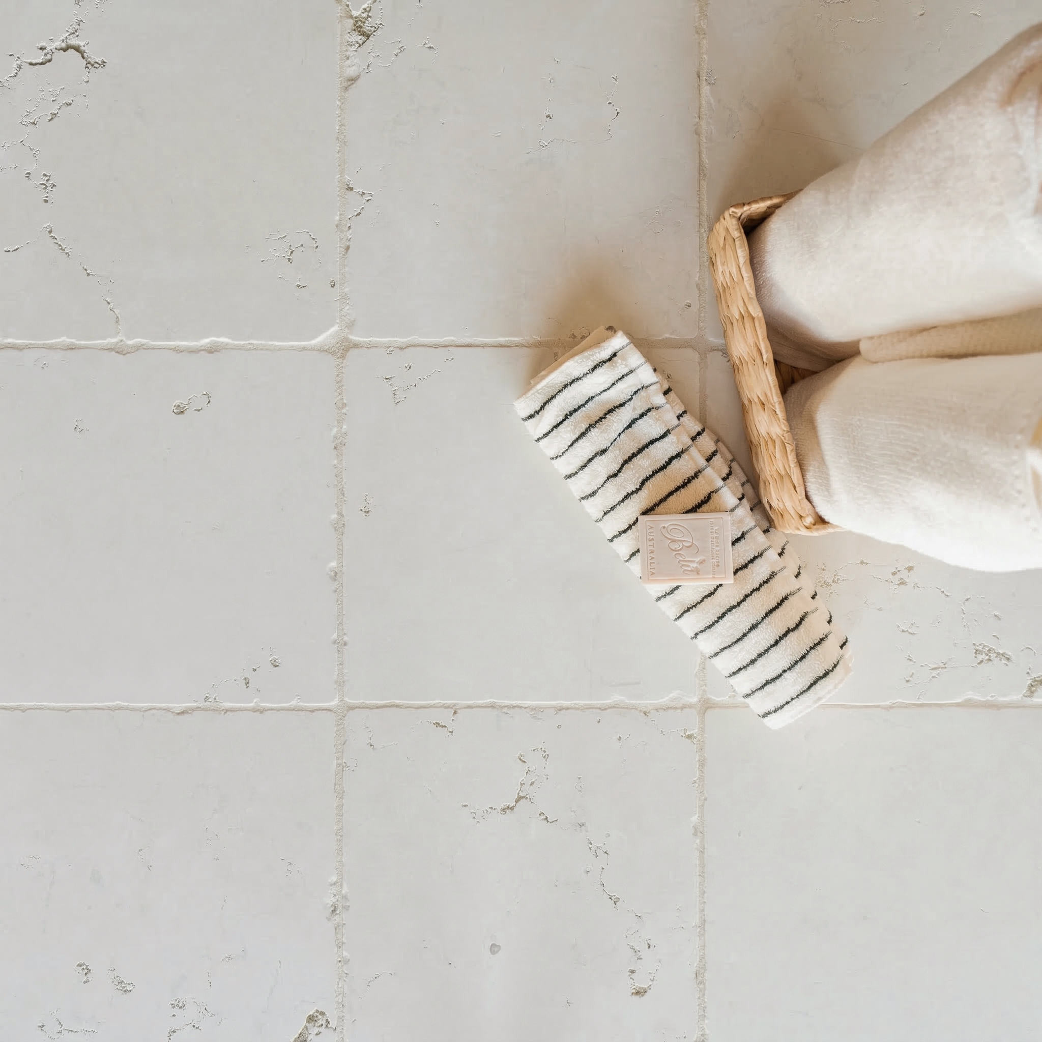 Tumbled off-white ceramic tiles with textured surface beside folded towels in a woven basket and striped cloth on floor