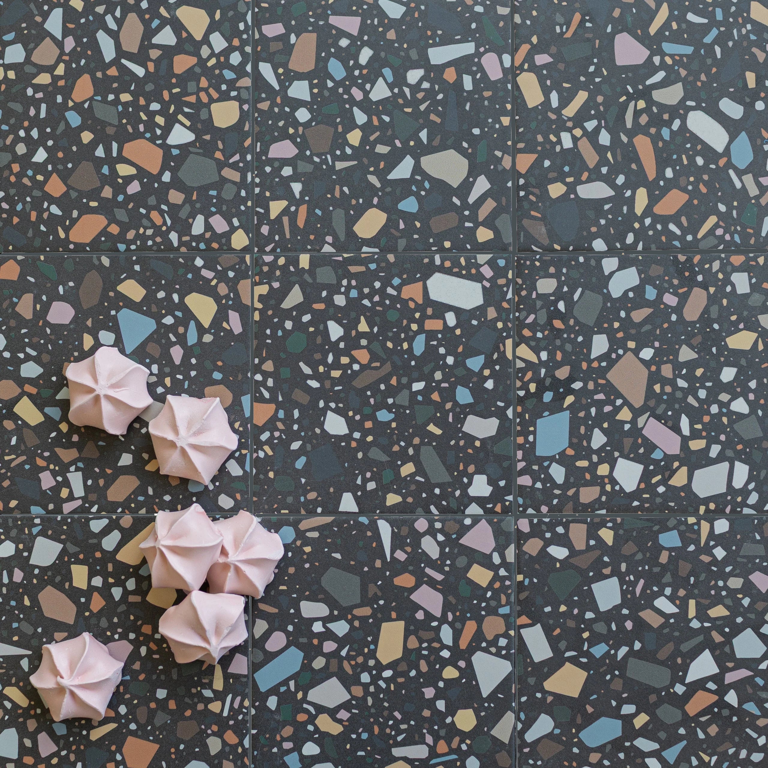Flat lay of dark terrazzo porcelain tile with scattered pastel stone chips and six pink meringue cookies clustered on surface