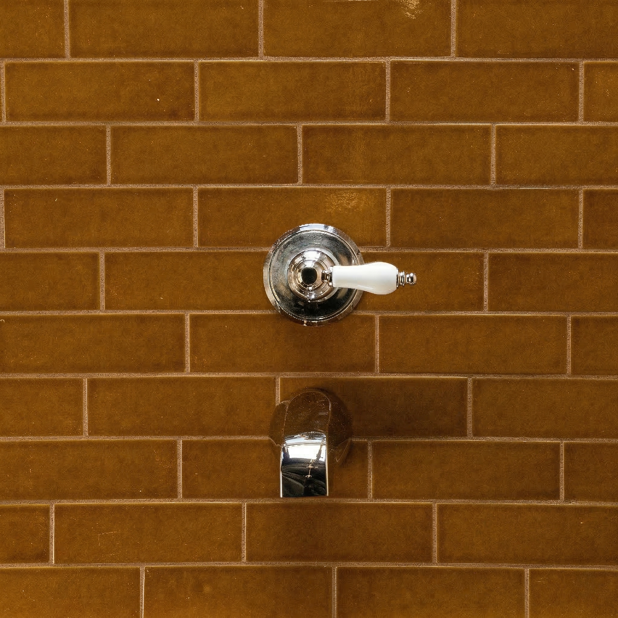 Glossy amber rectangular ceramic tiles arranged in a subway pattern with chrome faucet handle and spout mounted on the wall
