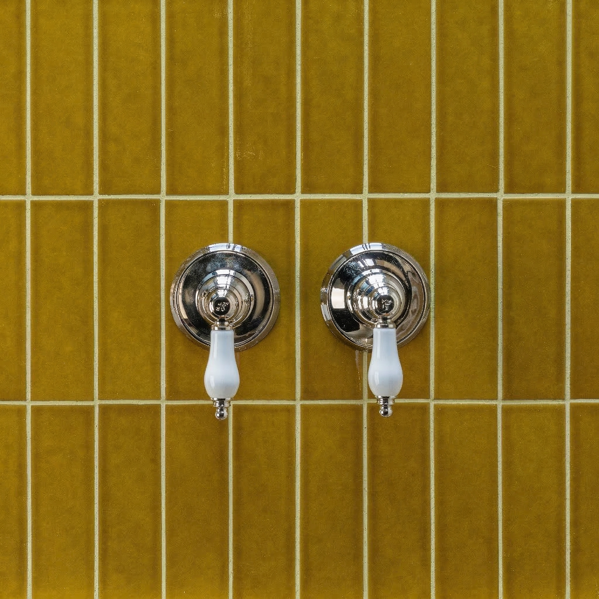 Glossy honey-colored ceramic tiles in a vertical grid with two vintage-style silver faucet handles and white levers
