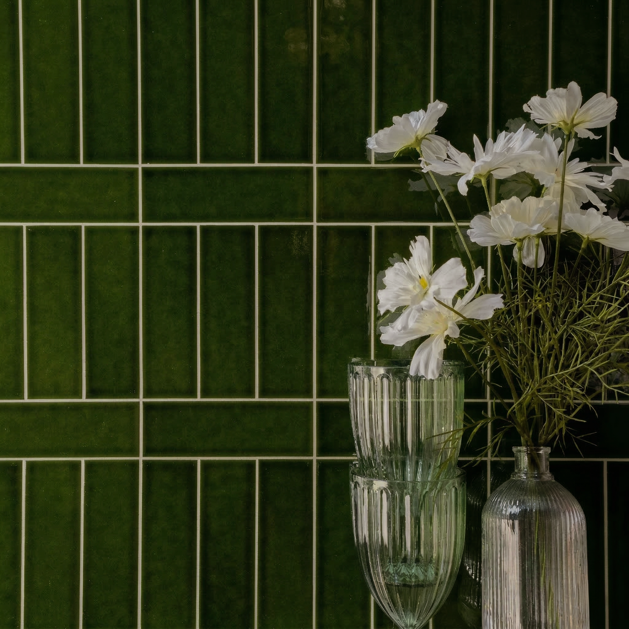 Glossy deep green ceramic tiles in a vertical and horizontal grid pattern with clear textured glass vases holding white flowers