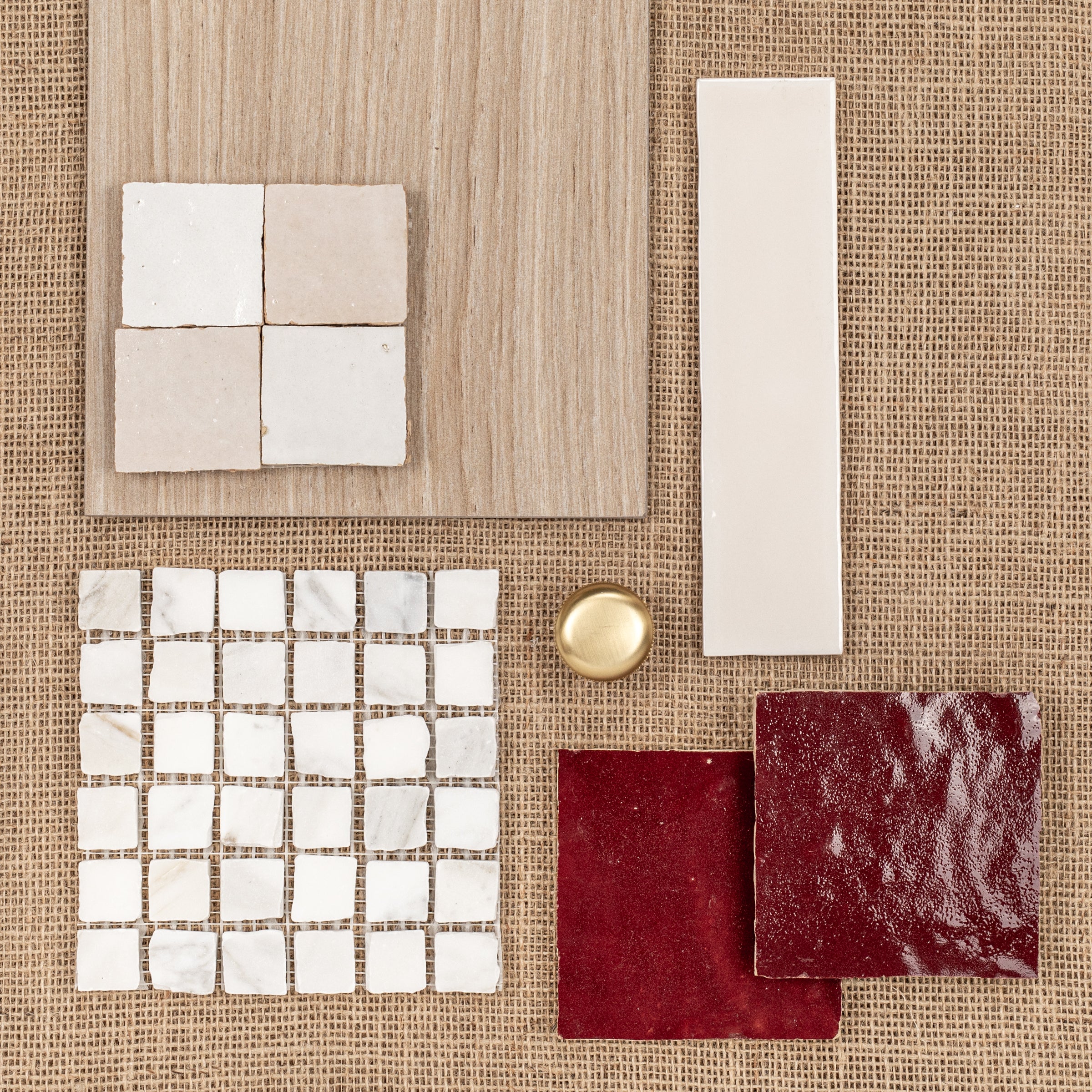 Assorted ceramic tile samples in neutral tones and deep red arranged on textured natural fabric with a brass knob nearby