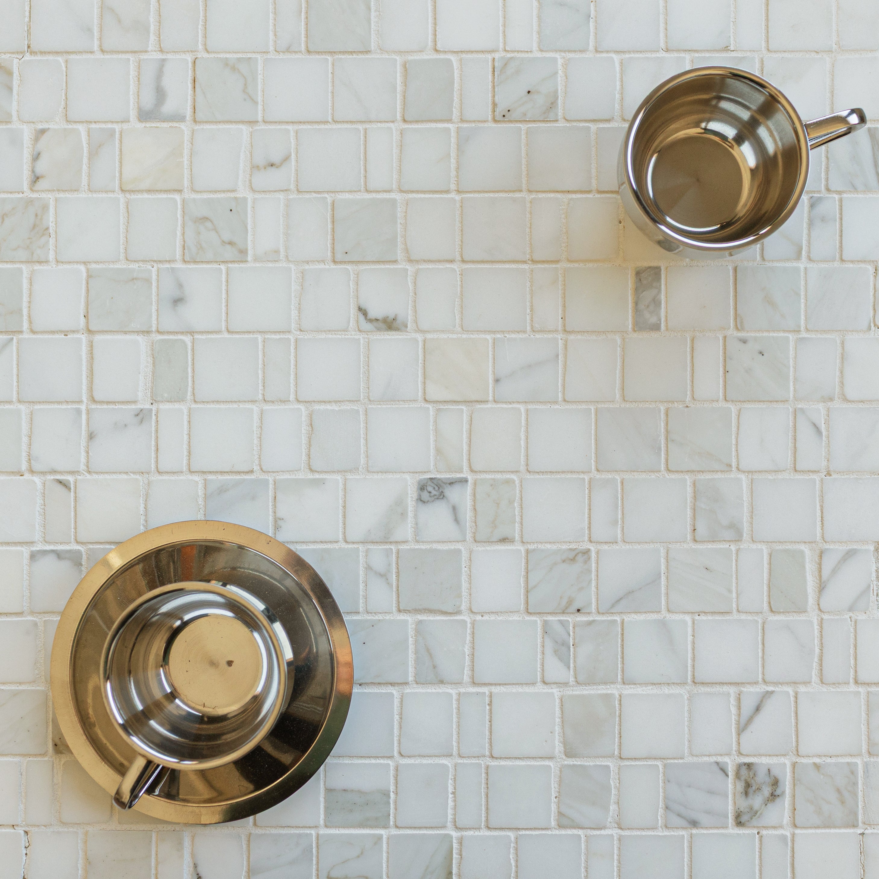 White and light gray marble mosaic tiles arranged in a subtle grid pattern with two stainless steel cups on top