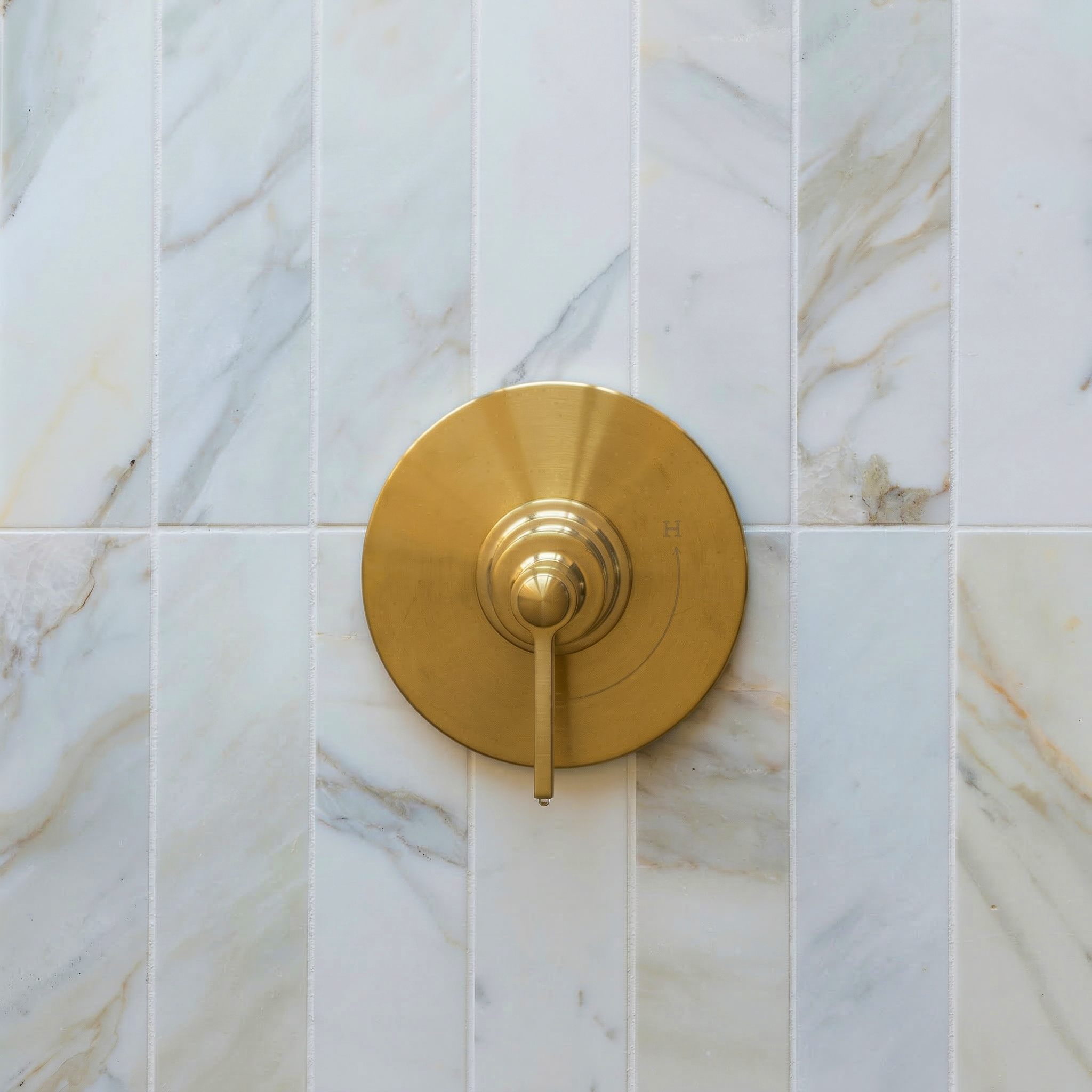 Brass shower control handle mounted on vertical white and gray veined marble tile wall with honed finish