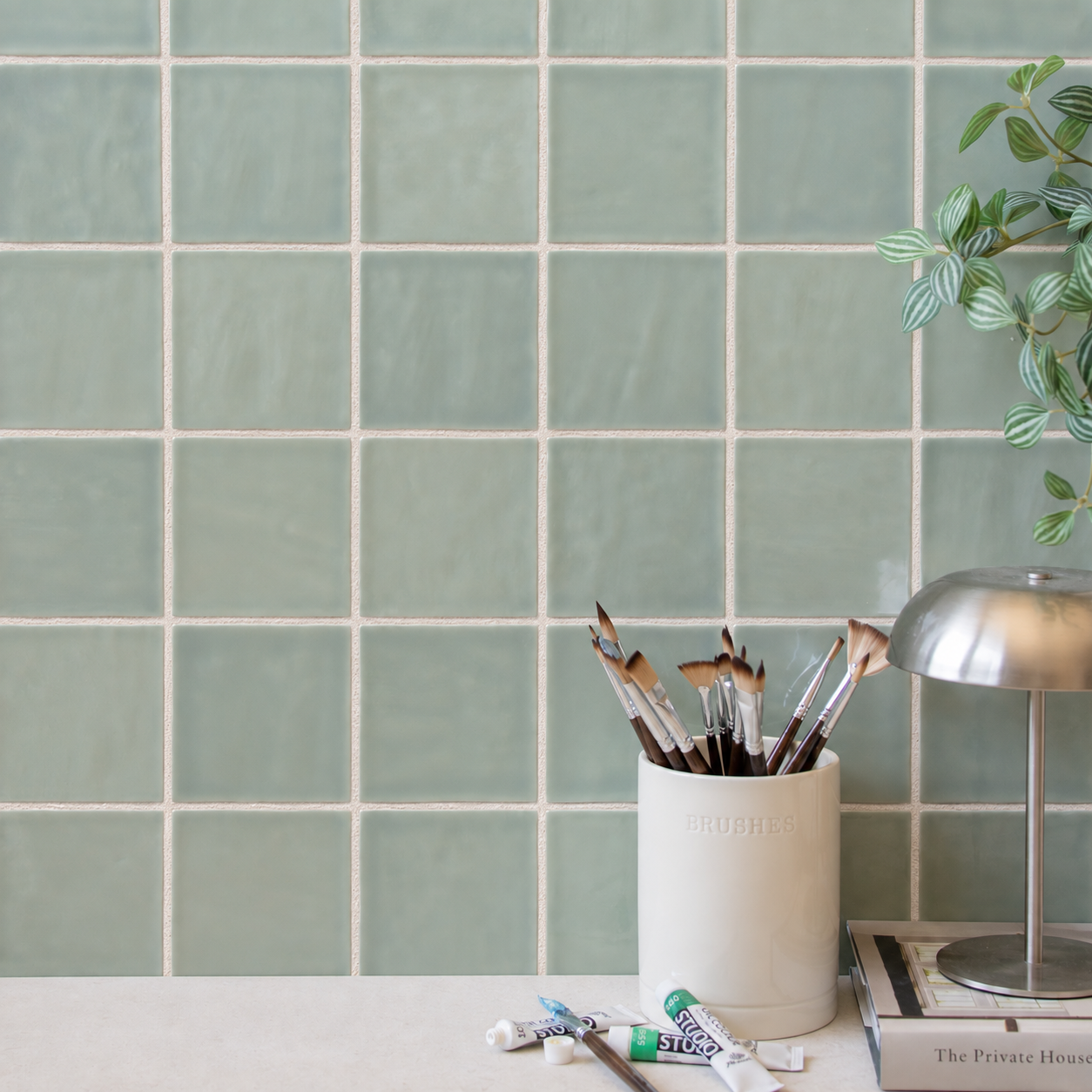 Green square tiled wall with paintbrushes desk lamp plant and art supplies