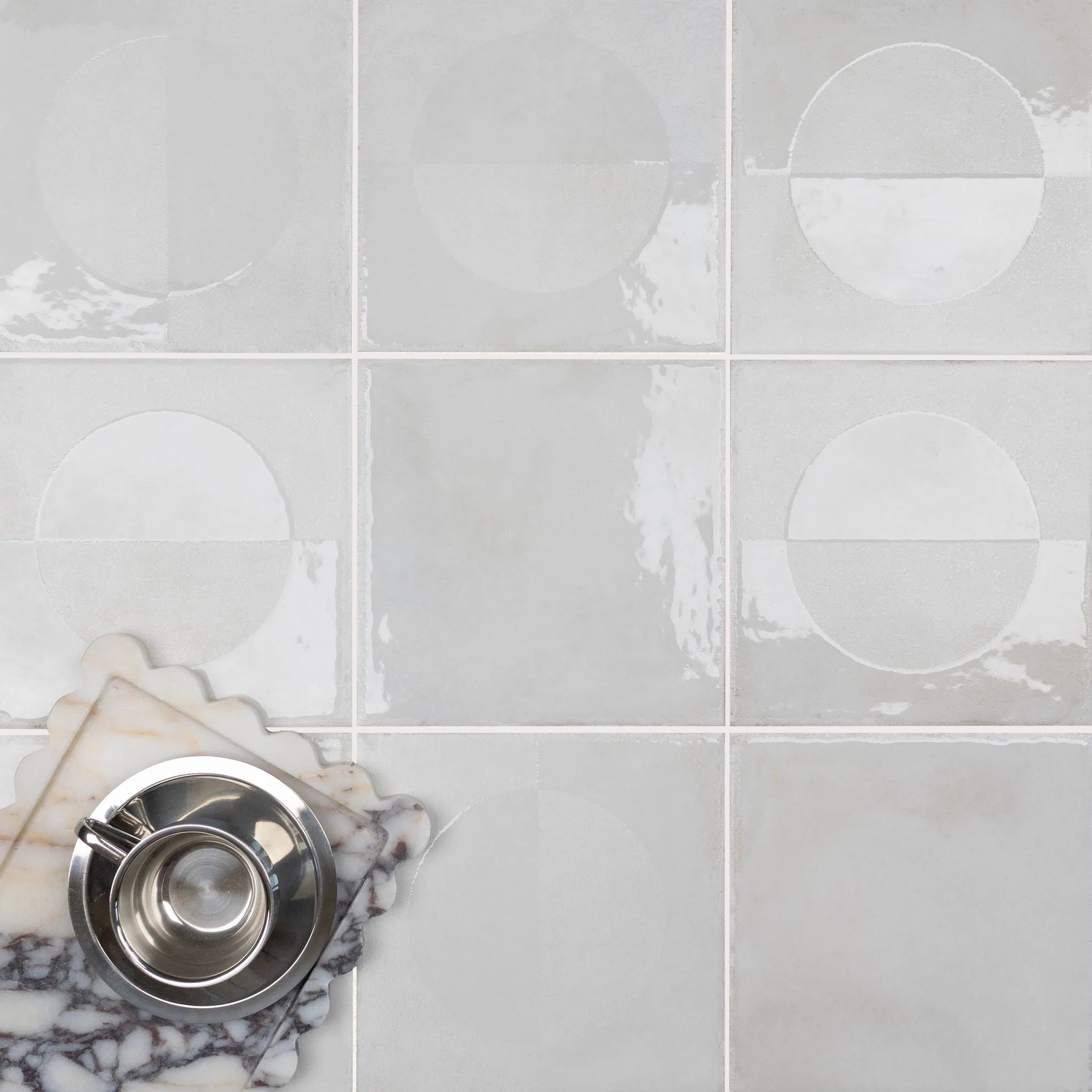 Glossy light gray porcelain tiles with subtle circular patterns and a stack of marble slabs holding a metallic cup and saucer