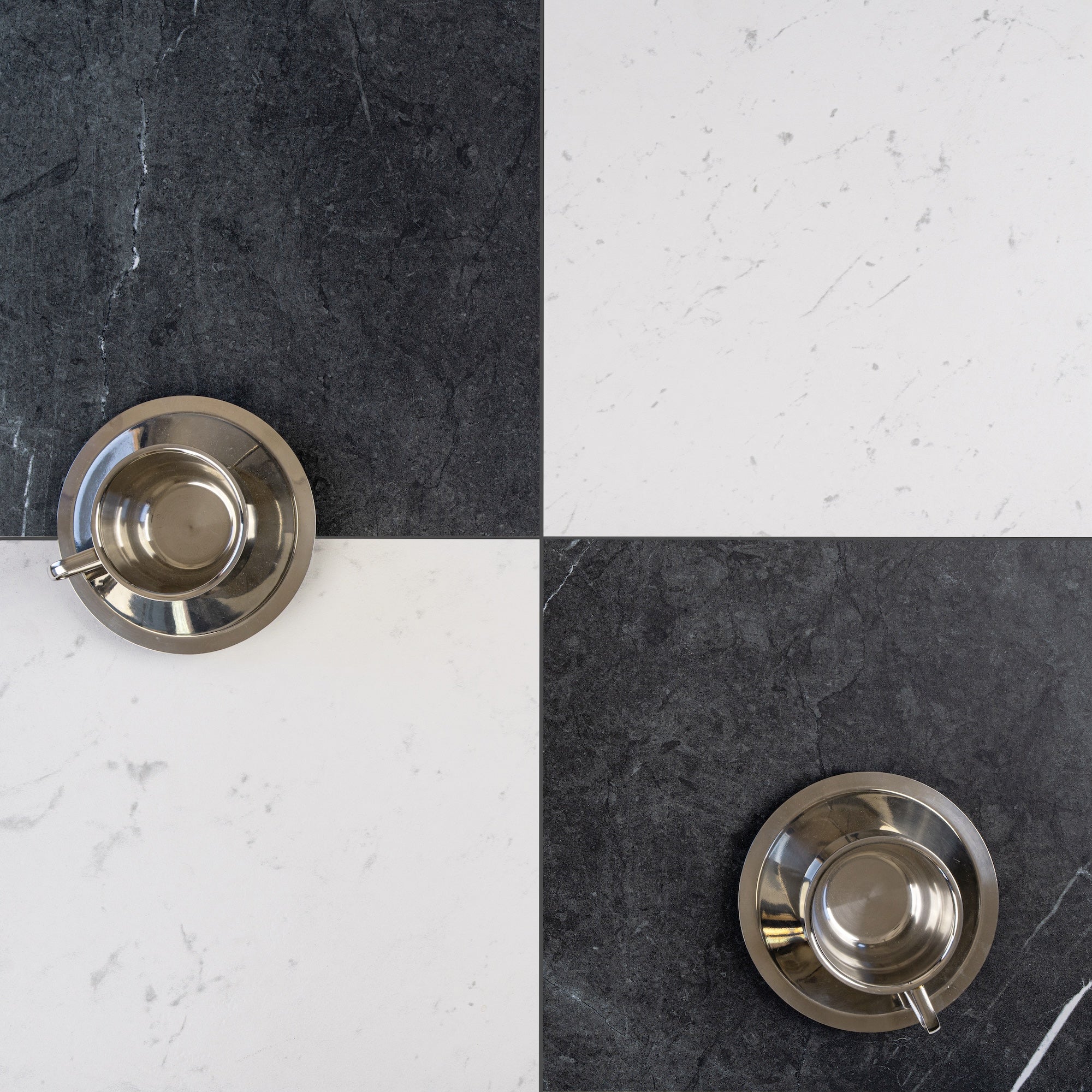 Two silver espresso cup and saucer sets on a black and white marble tiled surface arranged in a checkerboard pattern