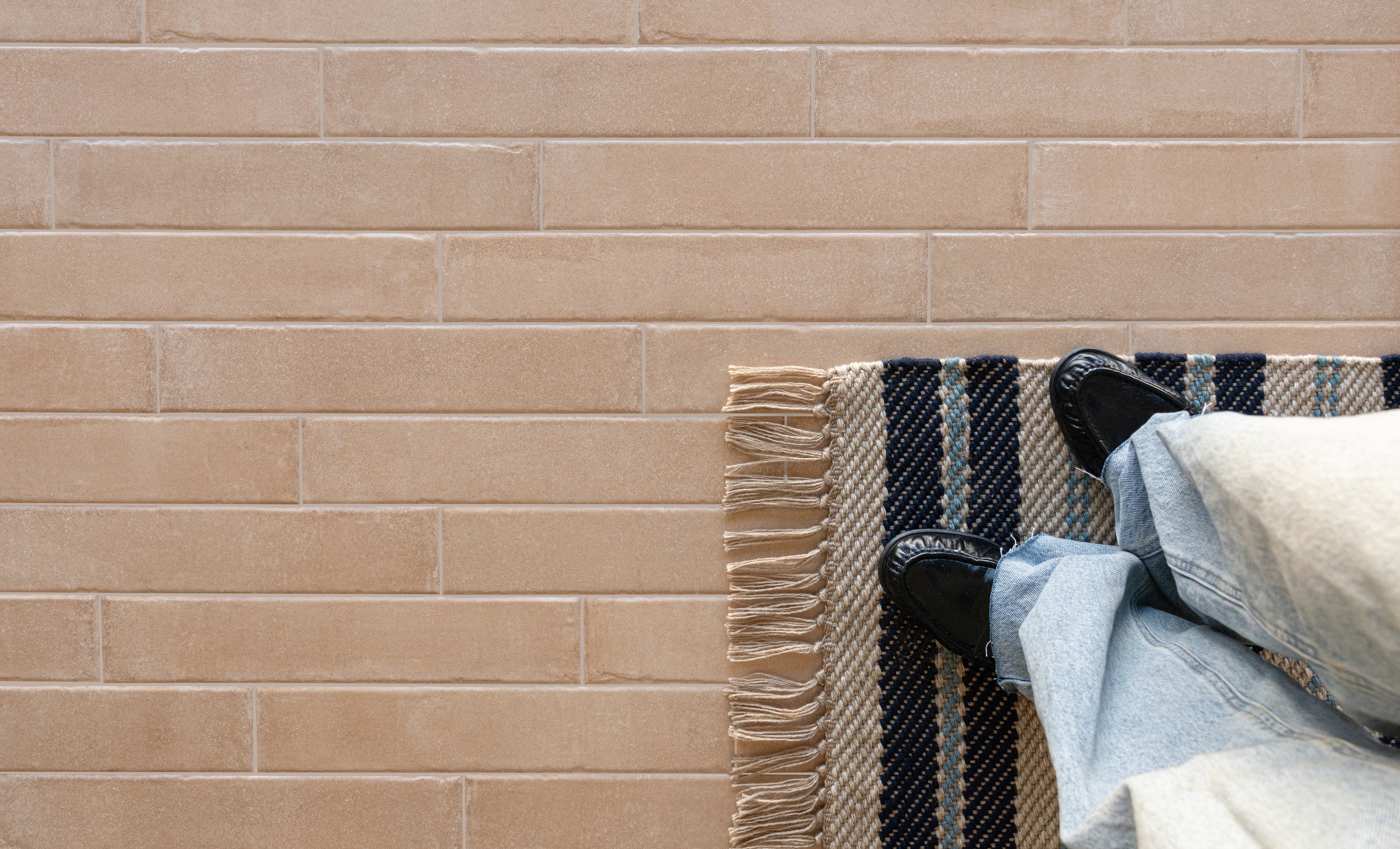 Soft beige rectangular tiles with subtle texture next to a striped fringed rug and person in denim and black shoes