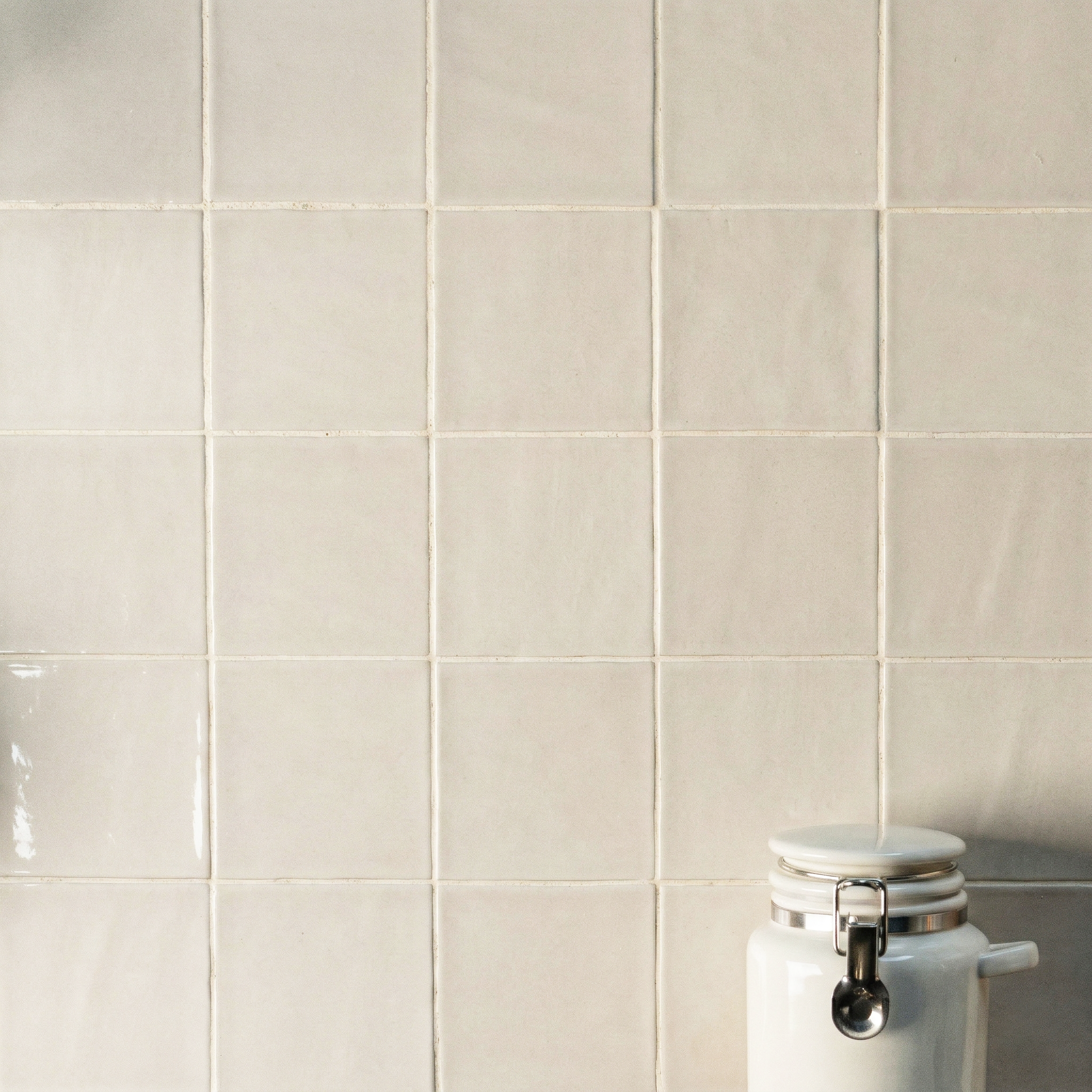 Matte-finish off-white ceramic tiles with subtle uneven texture arranged in a grid behind a white ceramic jar with metal clasp