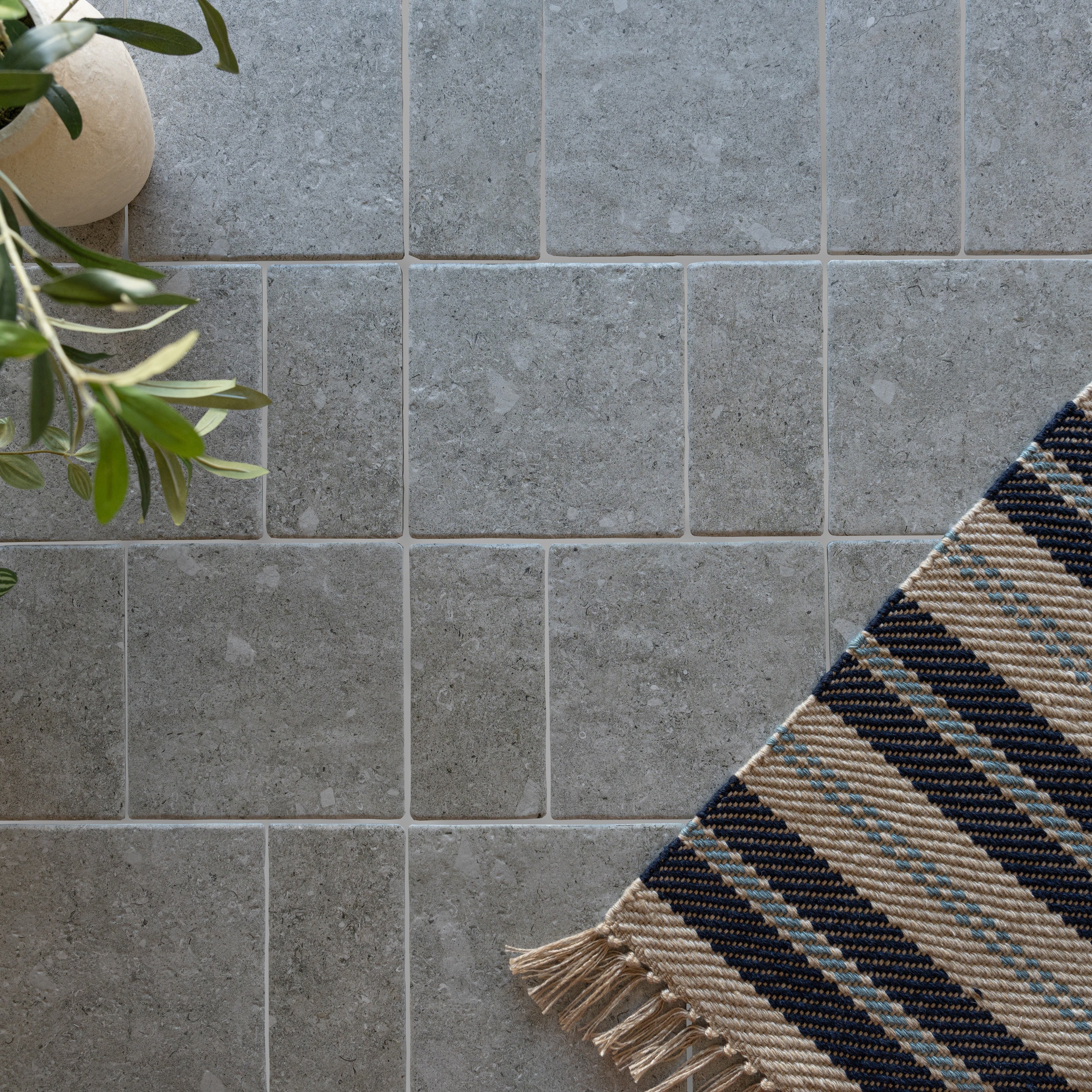 Gray porcelain tiles with subtle texture next to a leafy plant and a striped woven rug corner