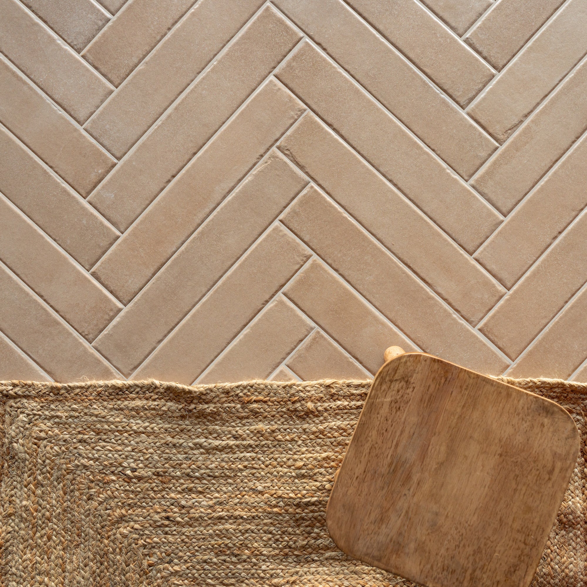 Beige large format tiles arranged in a herringbone pattern above a woven natural fiber rug with a wooden stool edge visible