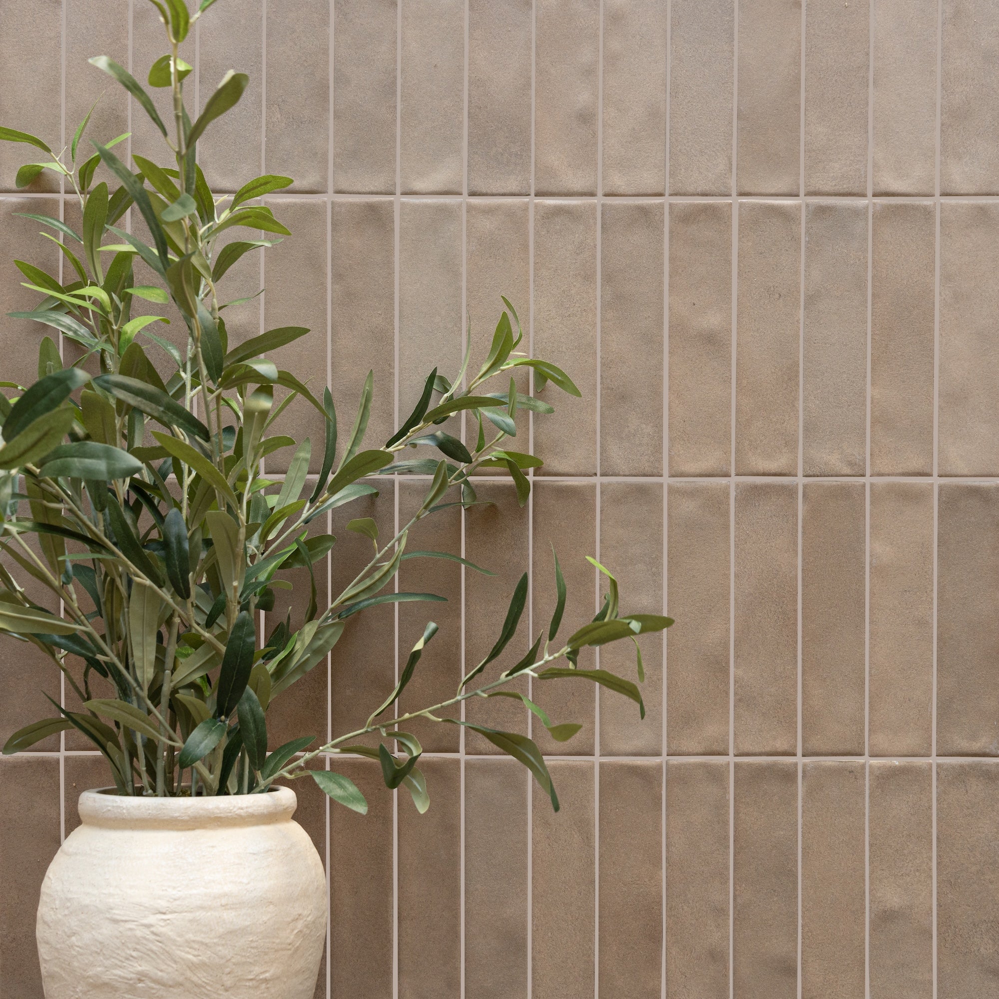 Matte beige ceramic tiles with subtle texture arranged vertically behind a white ceramic pot holding green leafy branches