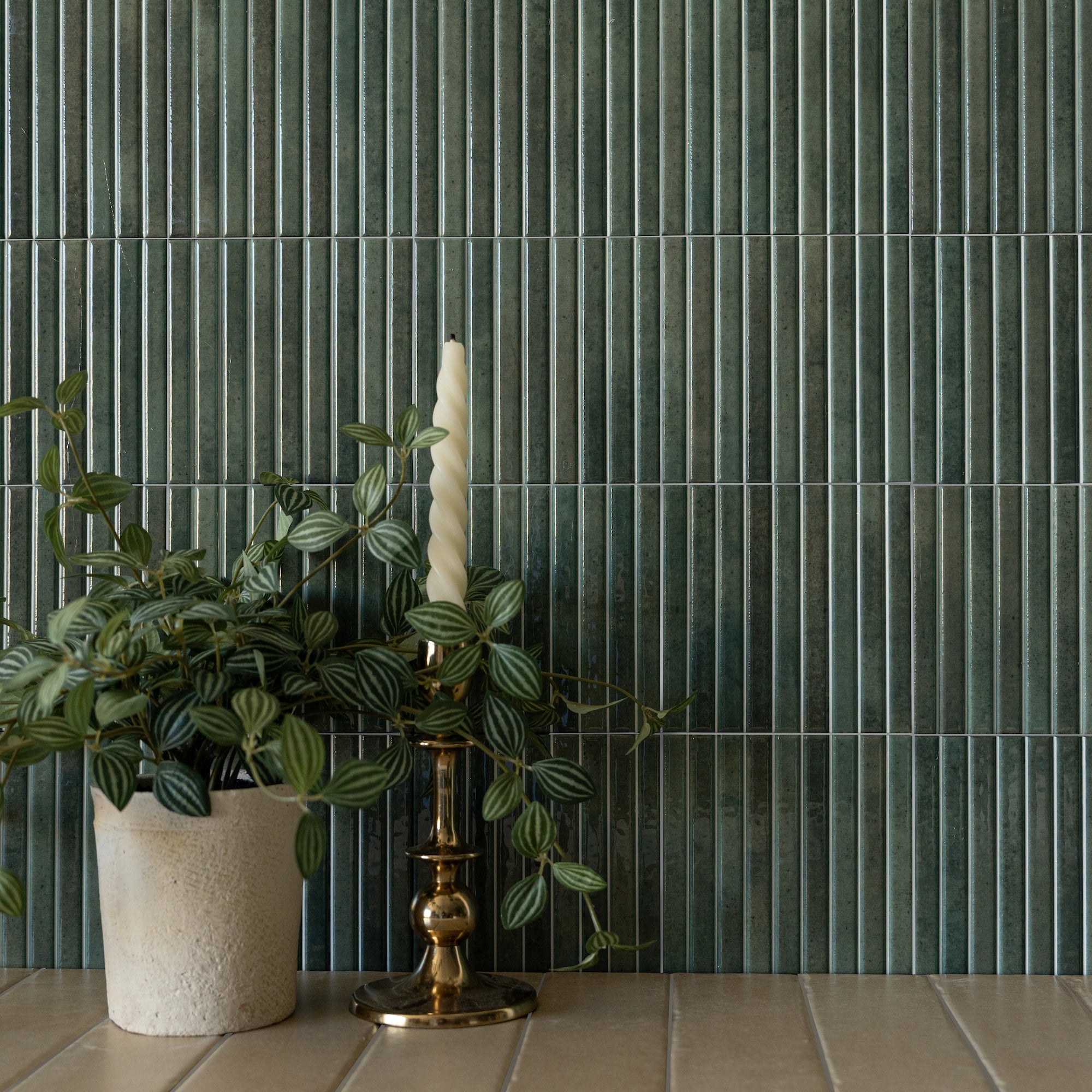 Glossy vertical ceramic tile wall in deep green with narrow white grout lines beside a potted plant and brass candle holder