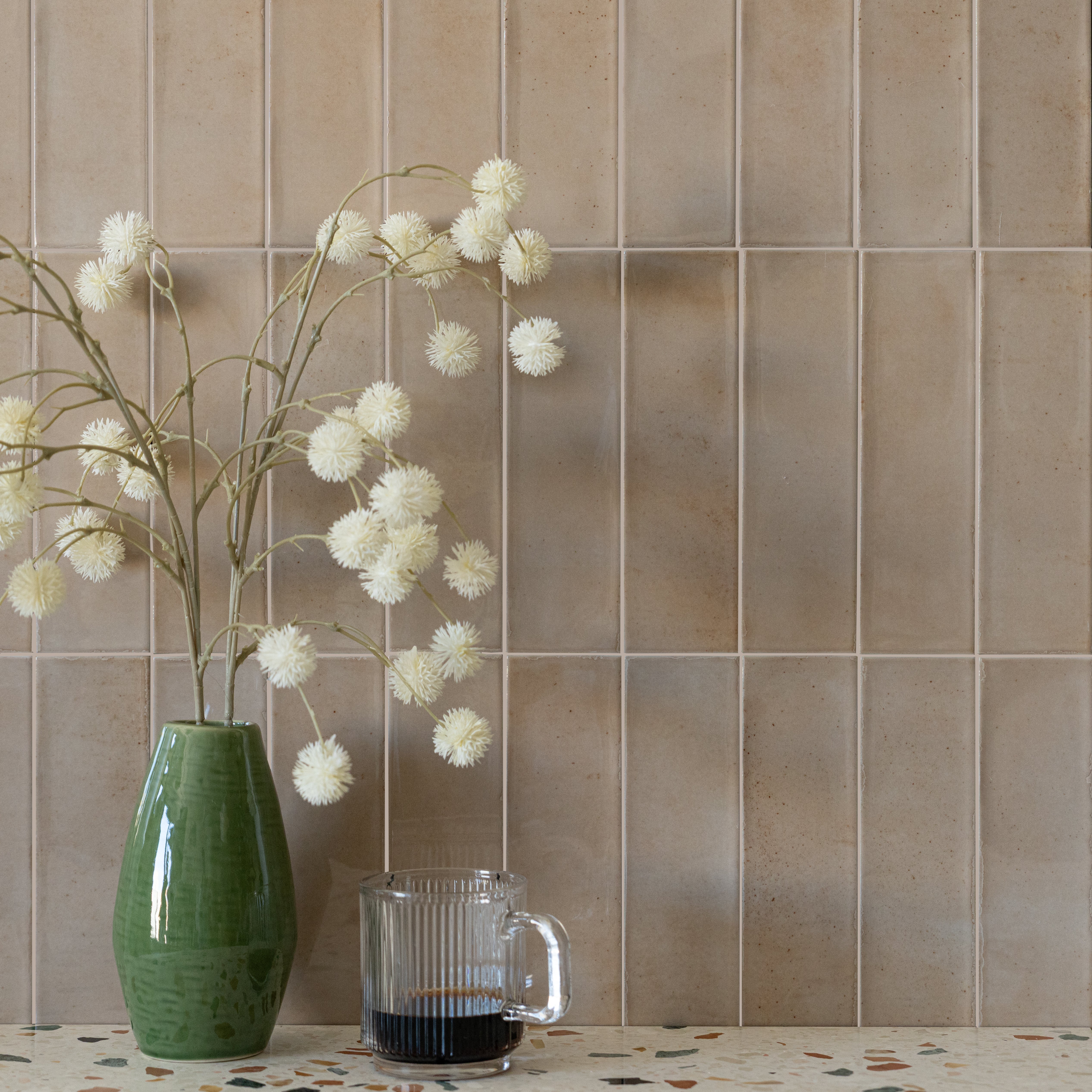 Green ceramic vase with white dried flowers and a glass cup in front of vertical beige ceramic tiles