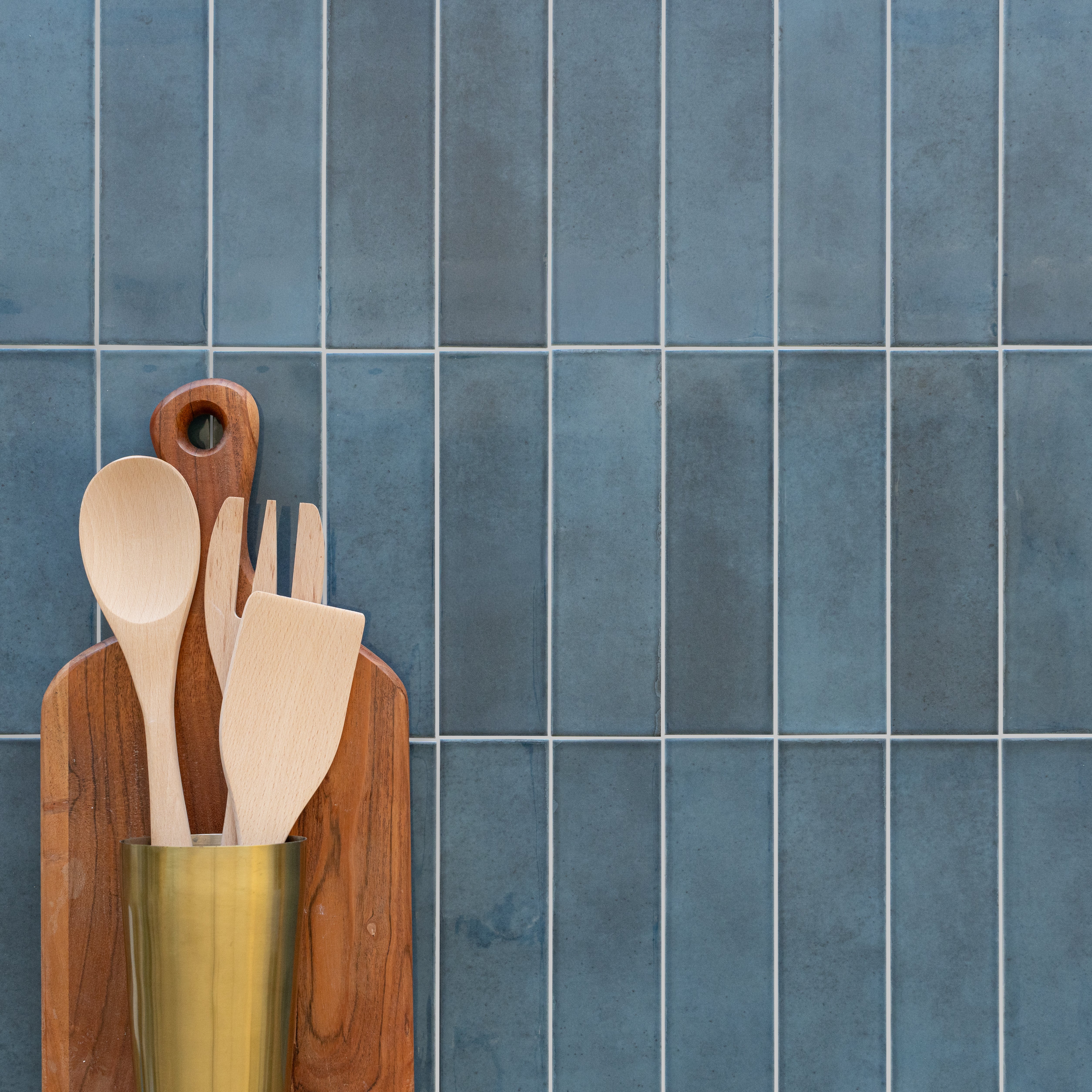 Vertical rectangular ceramic tiles in muted deep blue tones with wooden kitchen utensils in a brass holder and cutting boards