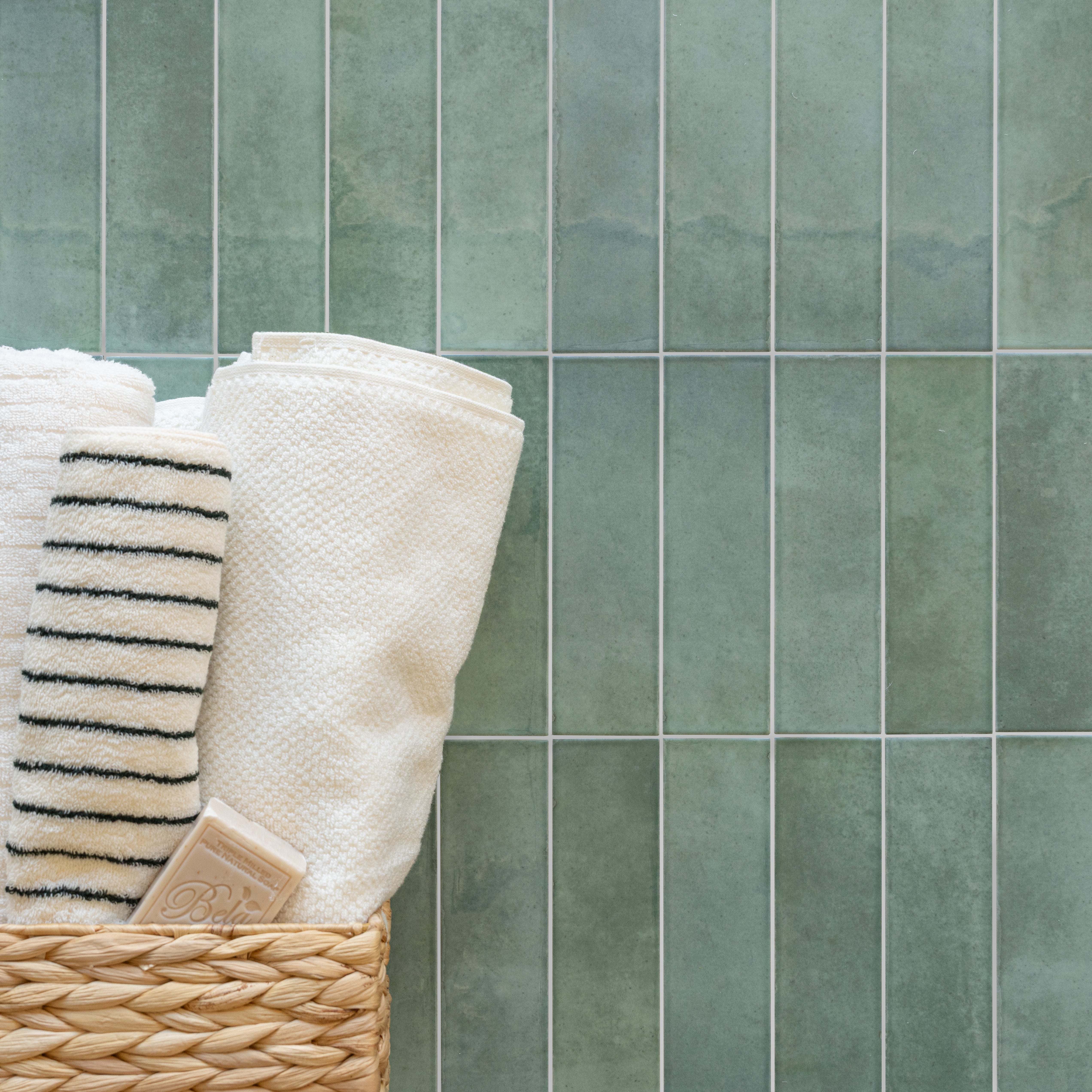 Matte ceramic tiles in soft muted sage green arranged vertically with a woven basket holding textured white towels and soap