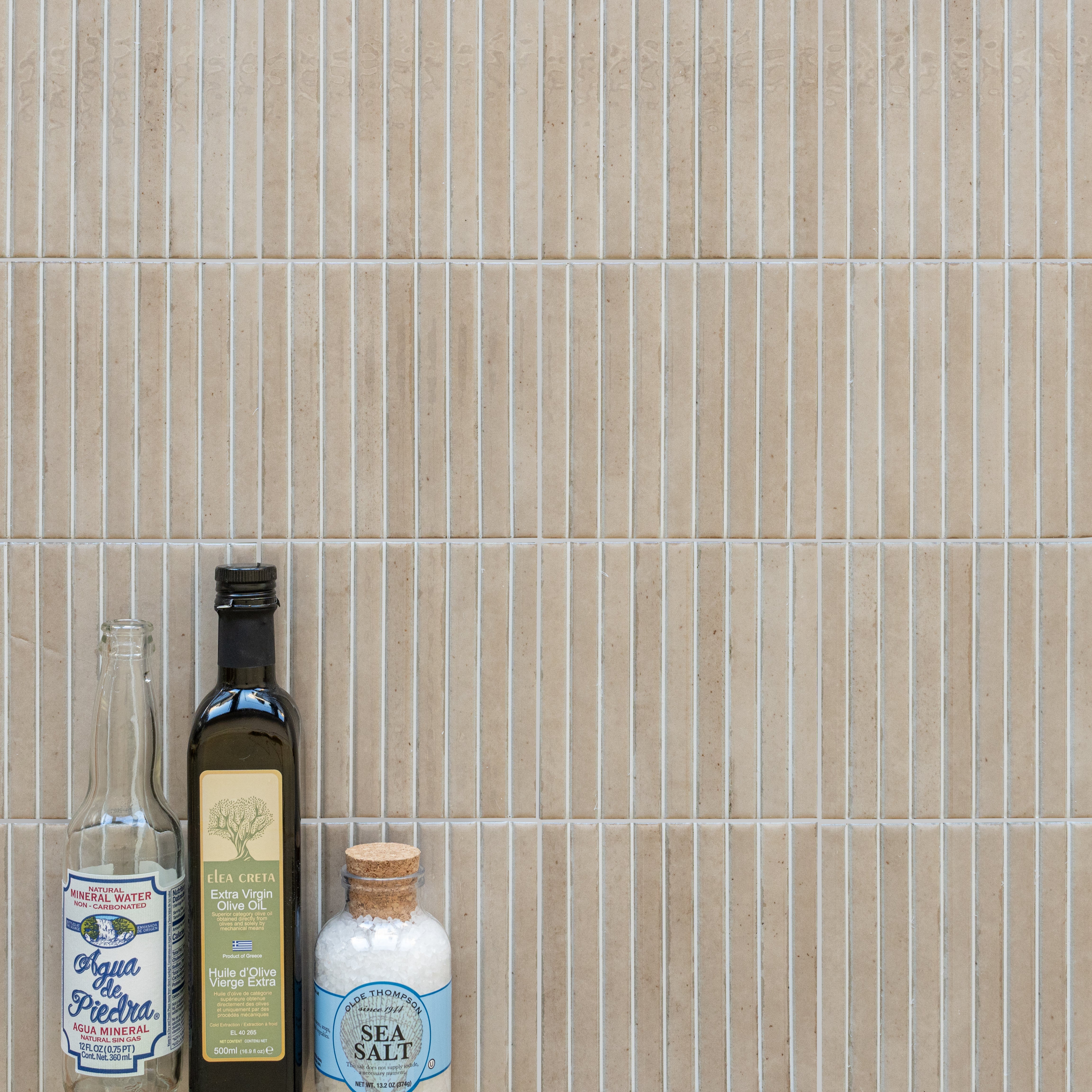 Vertical narrow beige ceramic tiles with thin white grout lines beside bottles of olive oil, sea salt, and mineral water