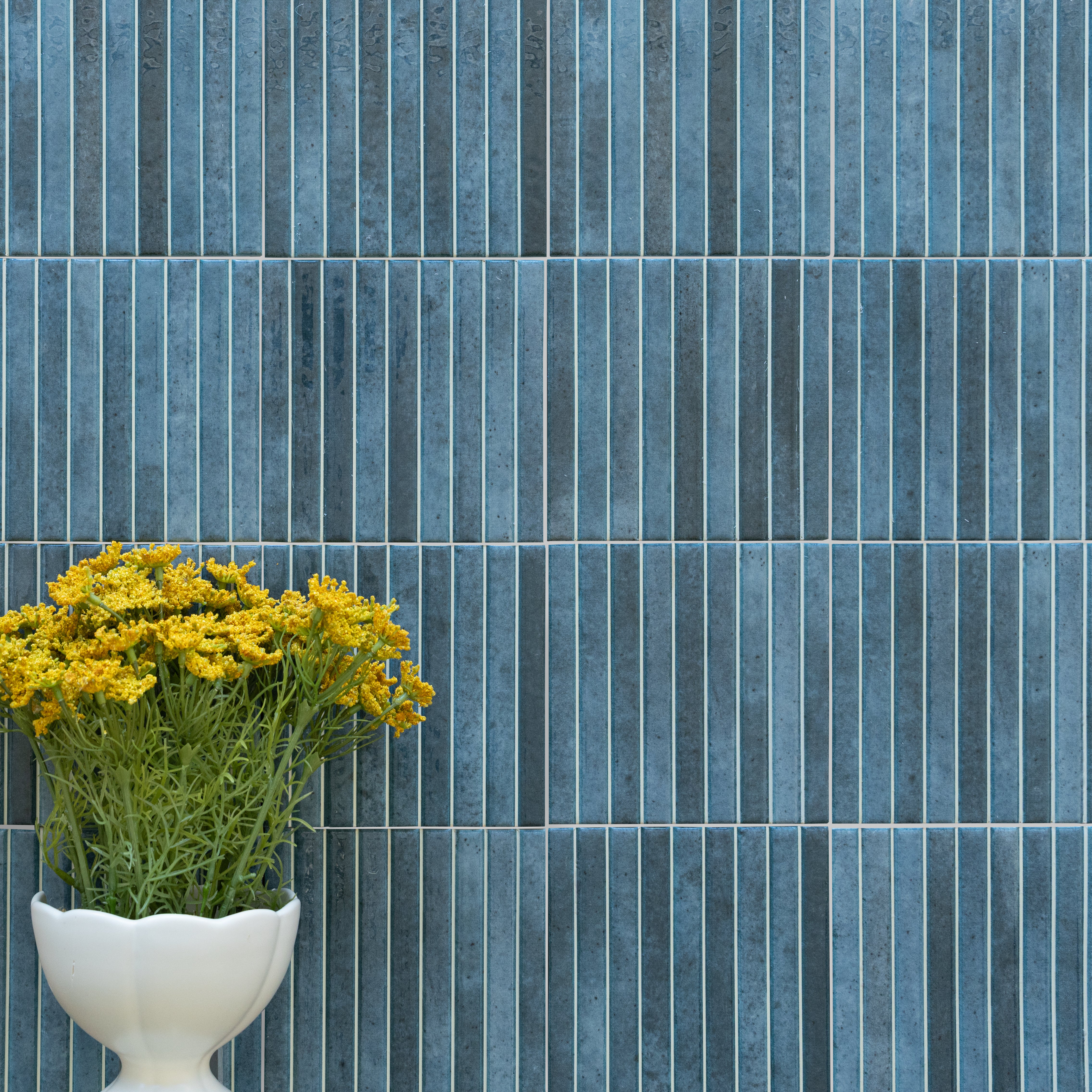 Vertical rectangular ceramic tiles in varied blue shades with subtle texture and narrow white grout lines behind yellow flowers in white vase
