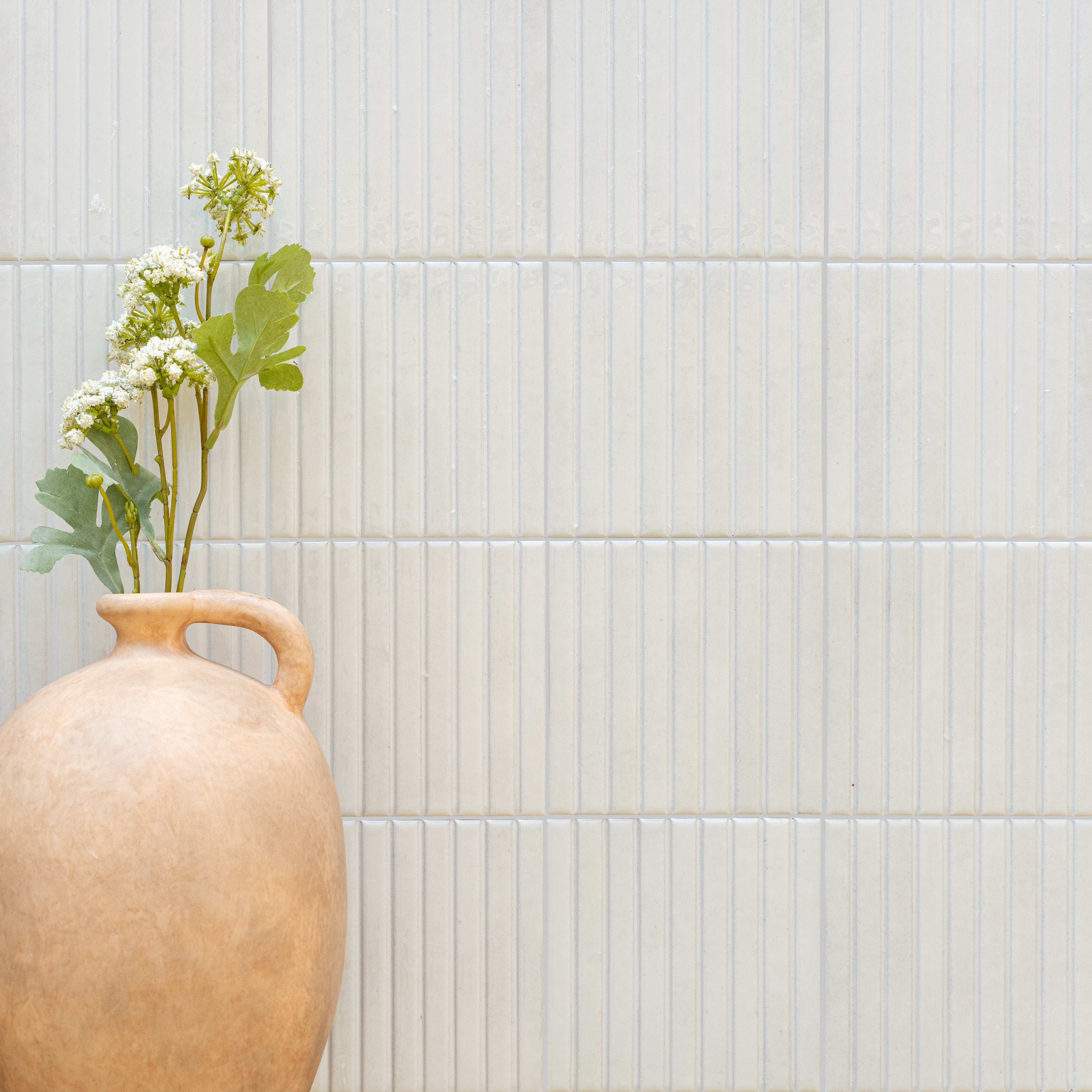 Matte white ceramic tiles with narrow vertical lines paired with a rustic earthenware vase holding green and white flowers