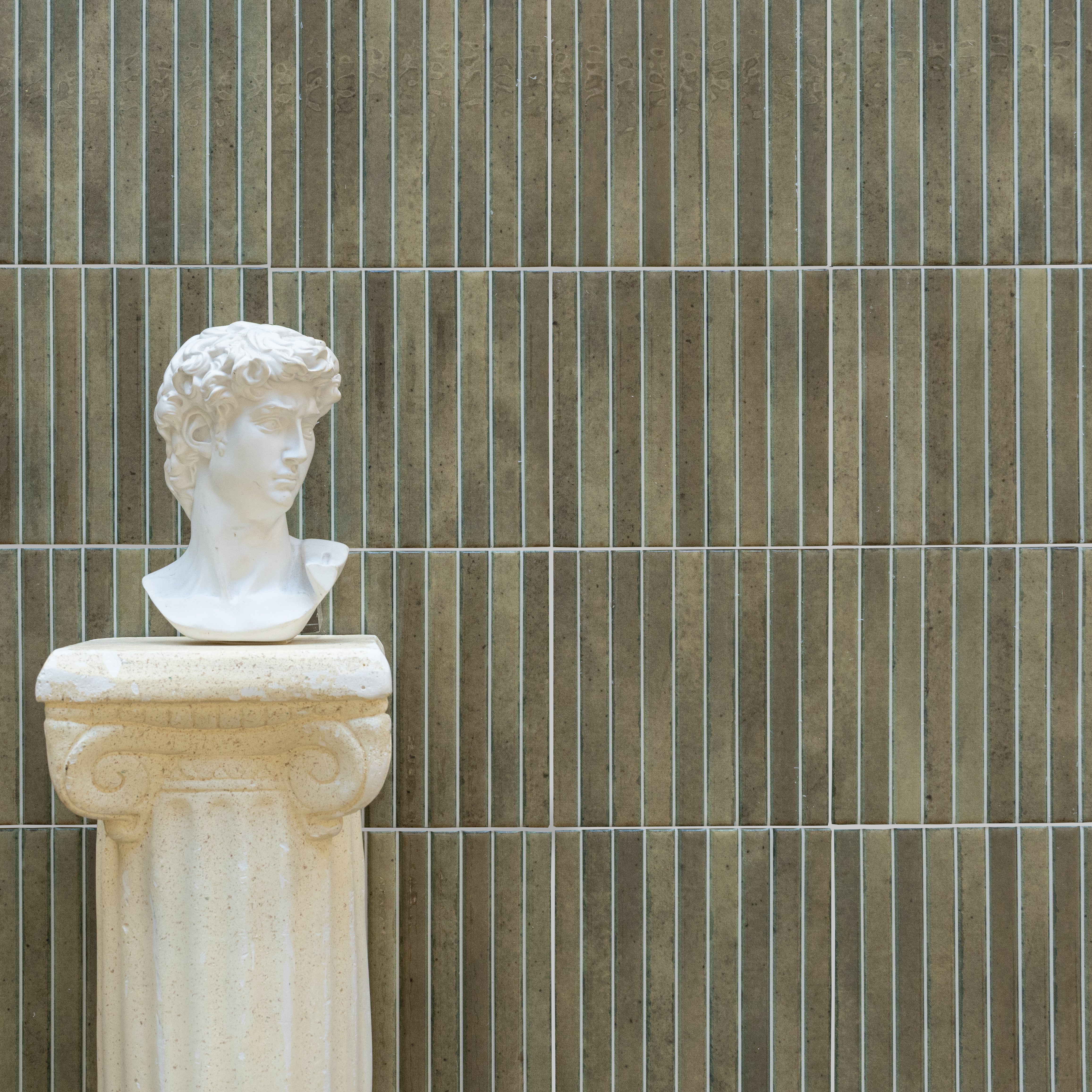 White classical bust sculpture displayed on a textured beige pedestal against vertically striped earthy brown tile wall