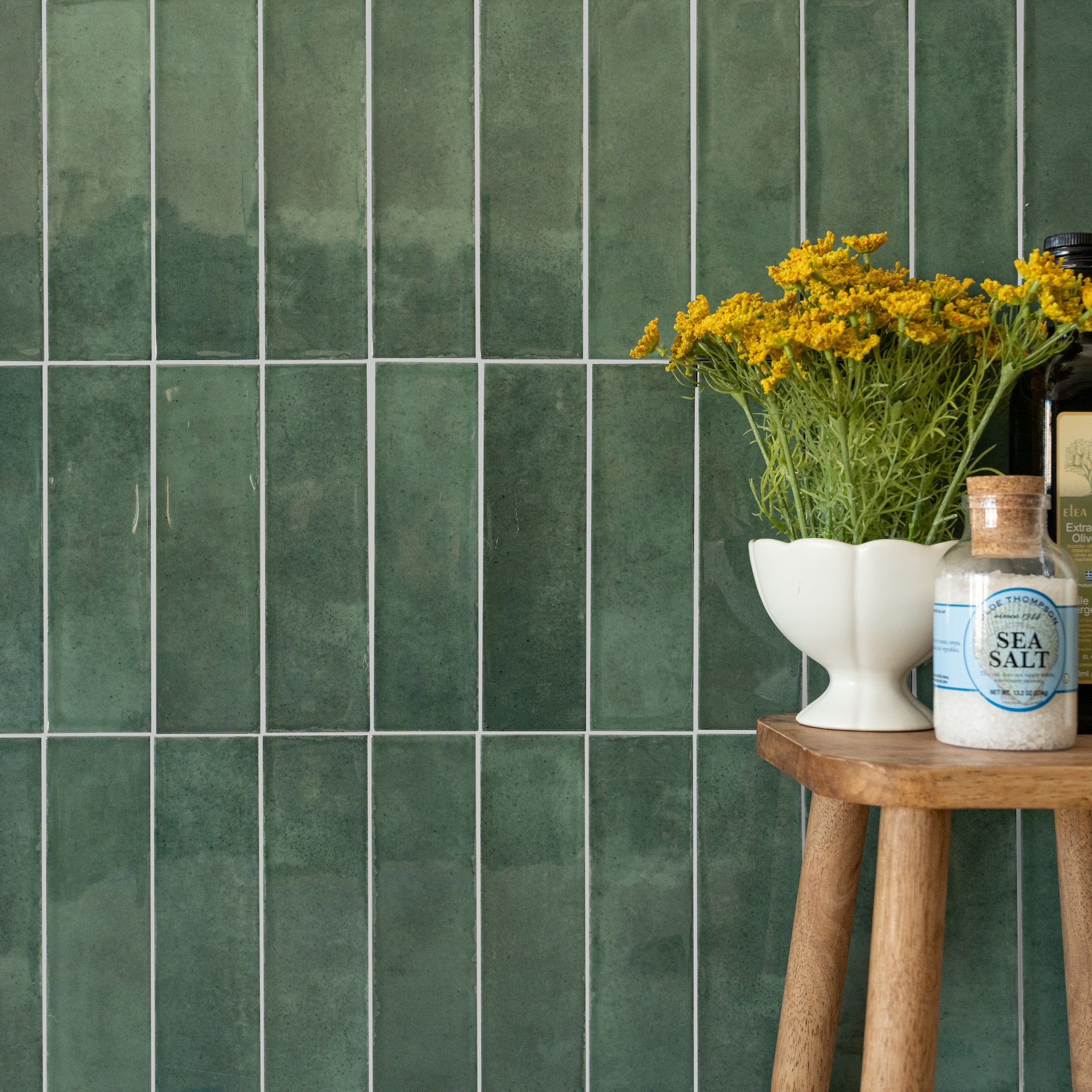 Glossy vertical green ceramic tiles with subtle texture beside a wooden stool holding yellow flowers and seasoning jars