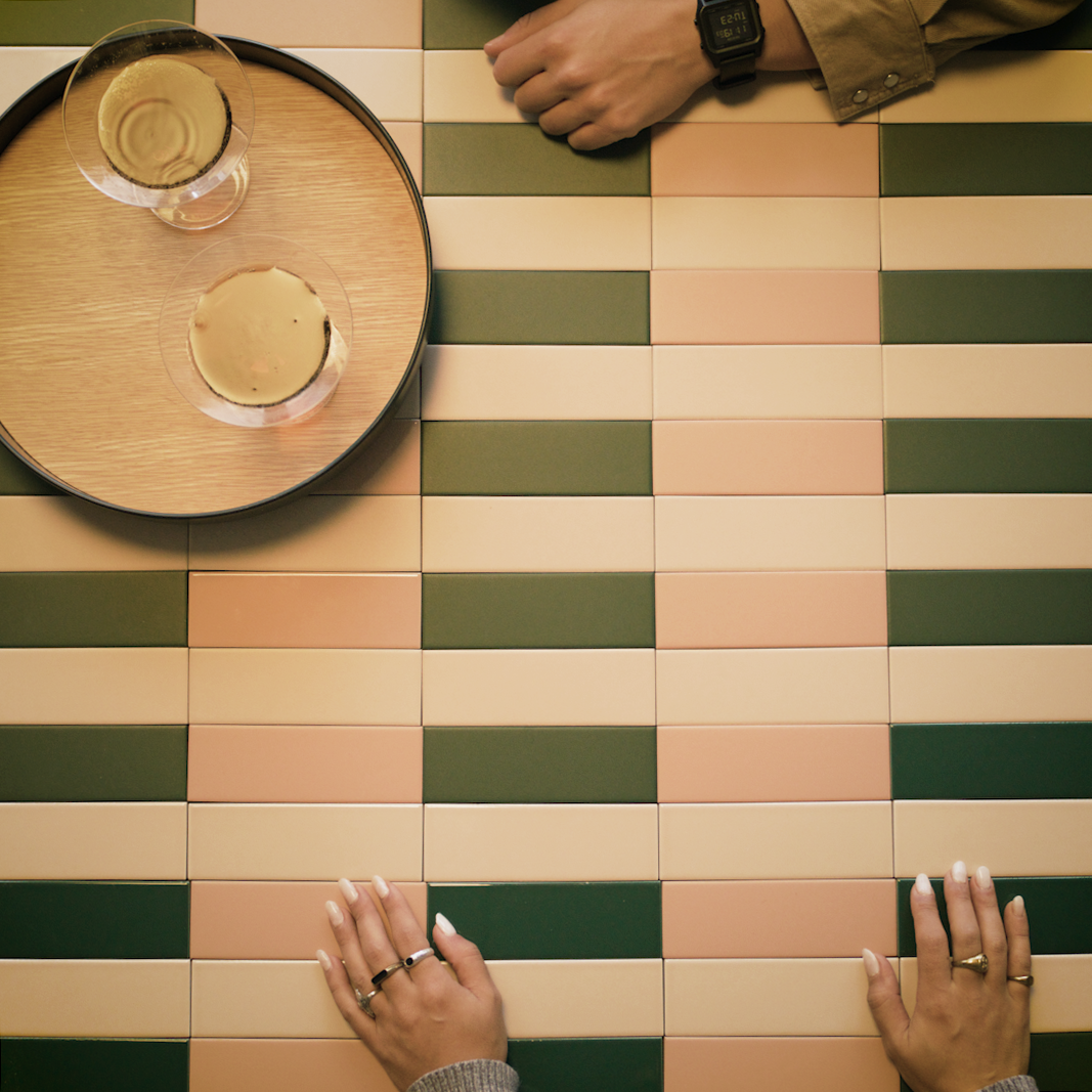 Overhead view of hands resting on rectangular ceramic tiles in green, peach, and cream colors with two drinks on a round wooden tray