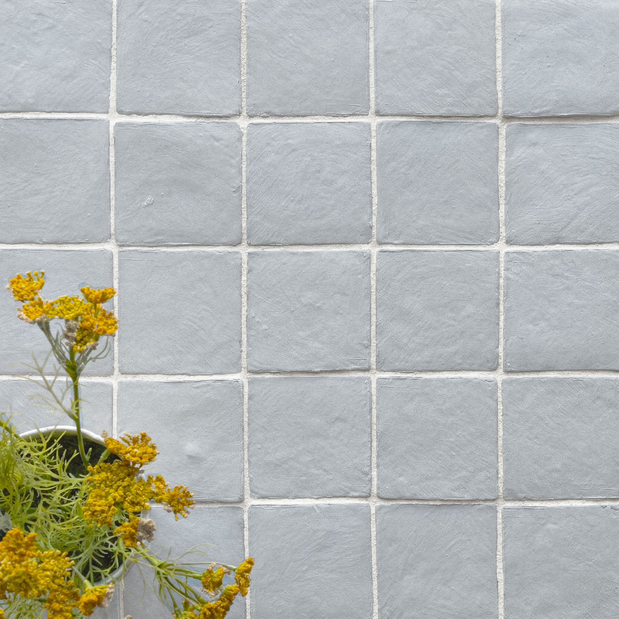 Matte blue ceramic tiles with subtle texture arranged in a grid beside yellow flowers in a small green pot