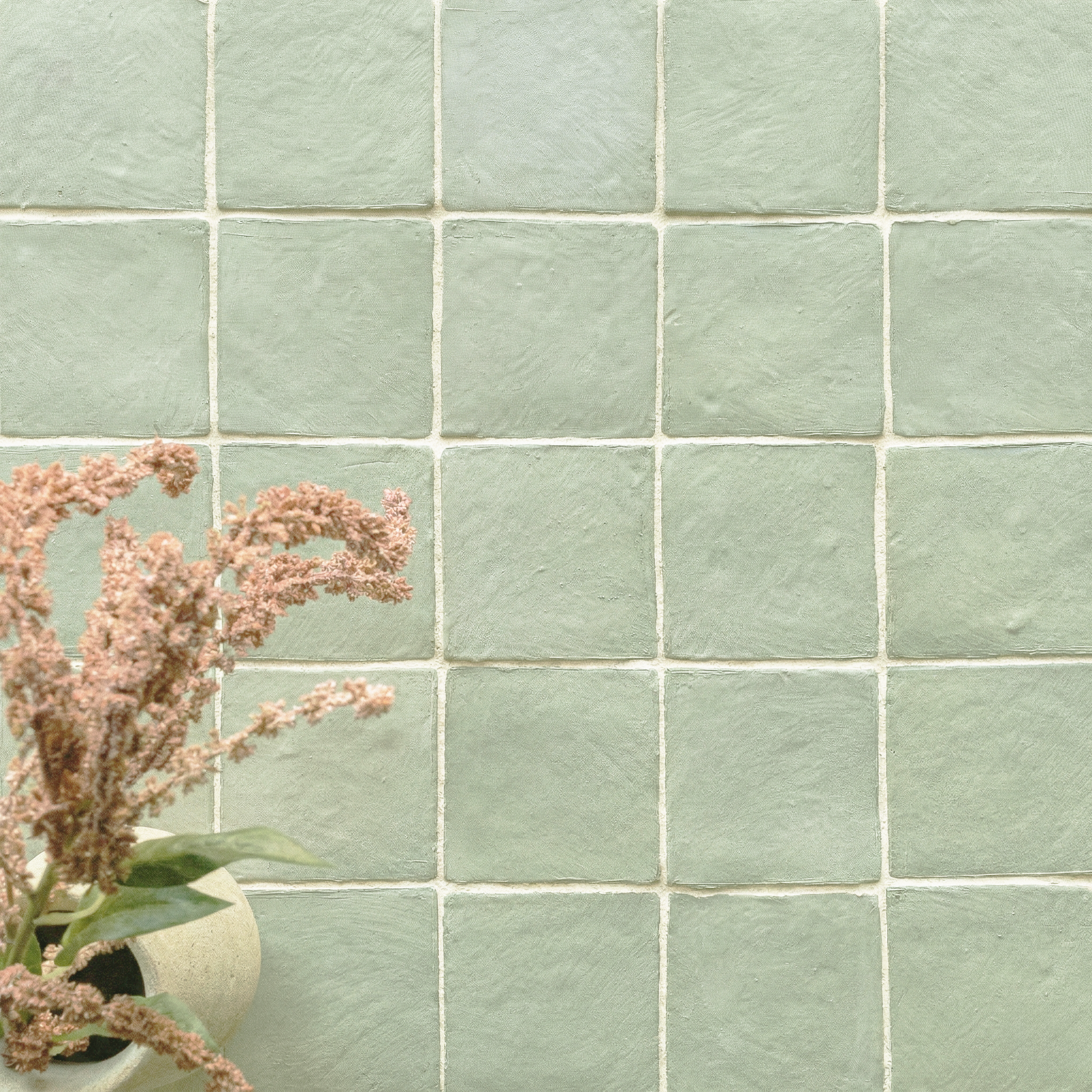 Soft green ceramic tiles with a handmade texture arranged in a grid beside a potted plant with pink flowers