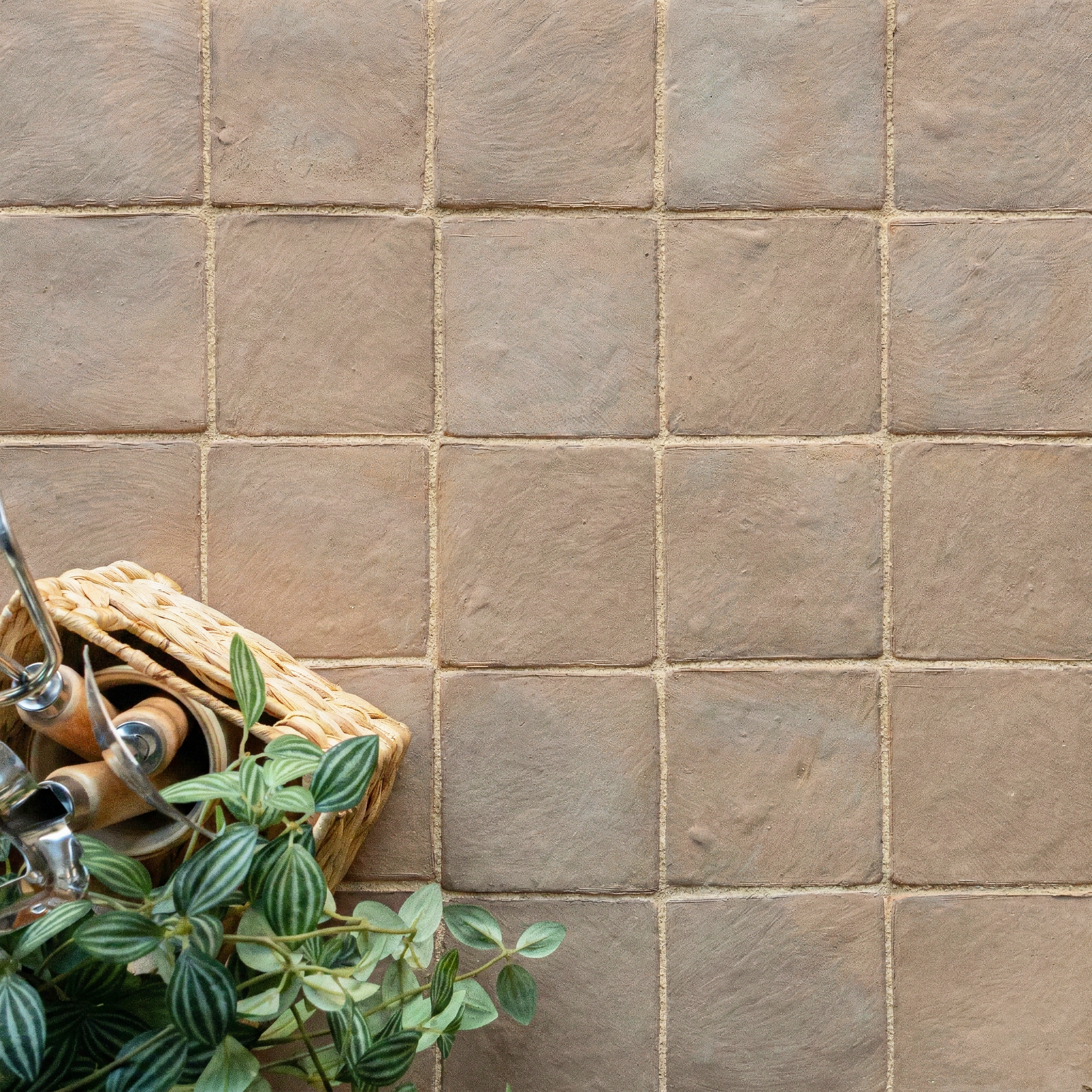 Textured sand-colored ceramic tiles arranged in a grid with a basket and leafy green plant on the corner
