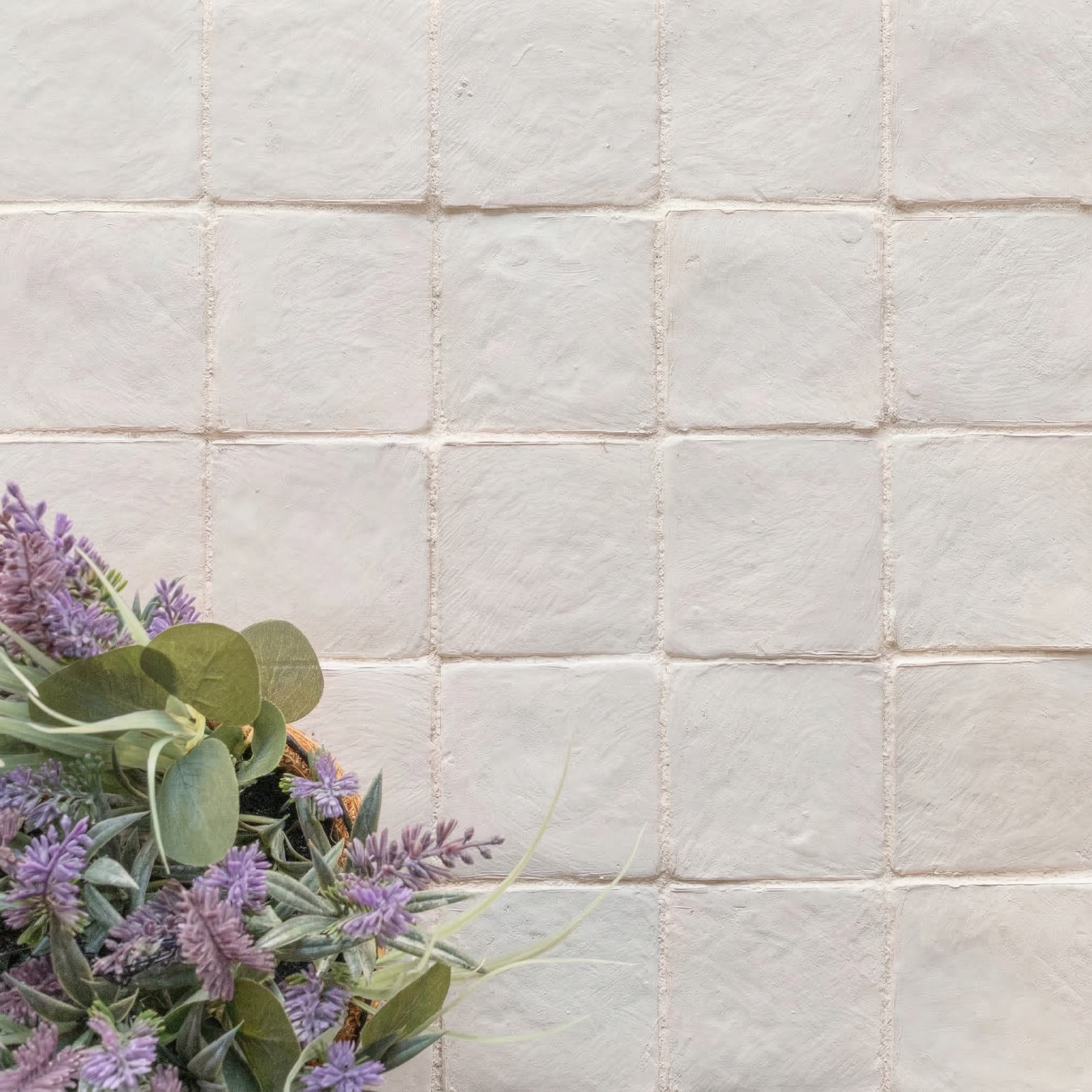 Off-white textured ceramic tiles arranged in a grid with a cluster of purple flowers and greenery at the corner