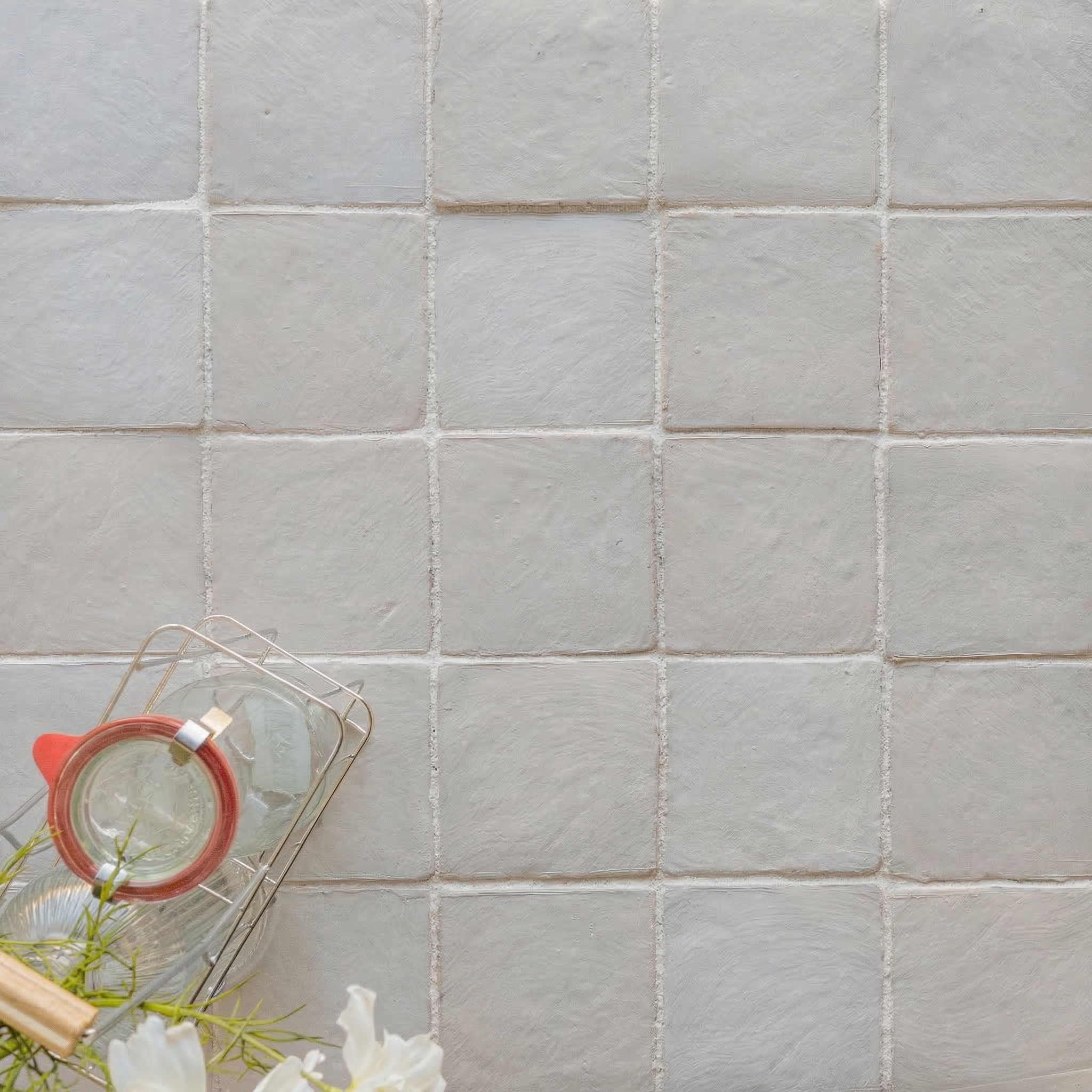 Soft pewter ceramic tiles with subtle texture arranged in a grid beside a glass teapot and white flowers on light surface