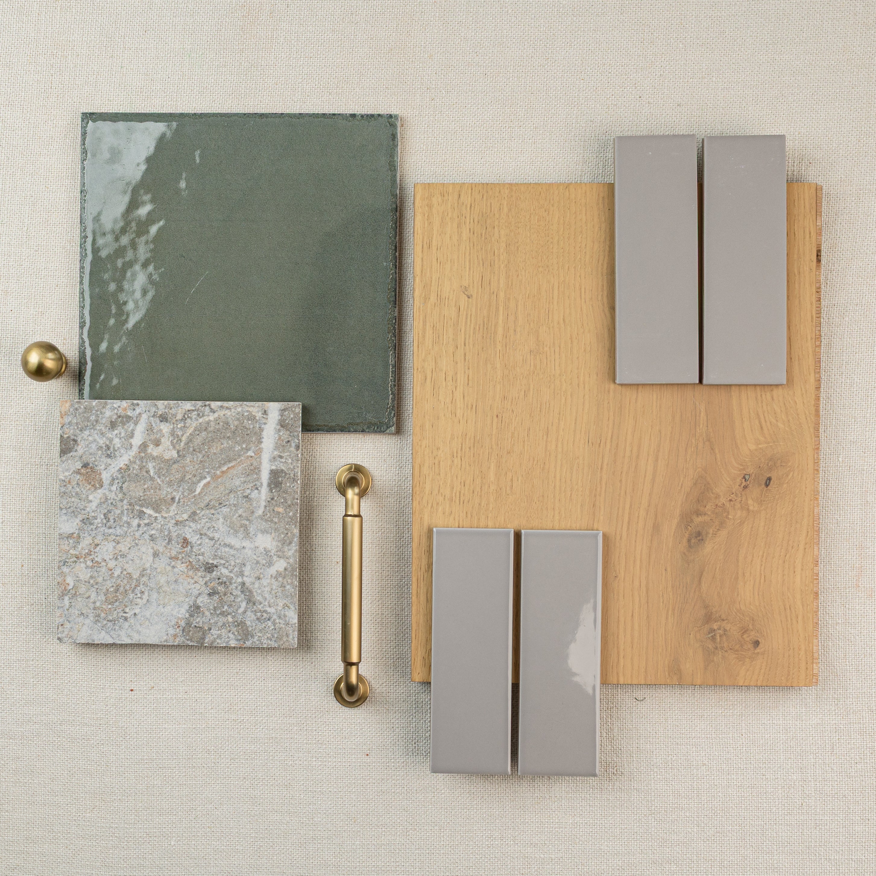 Green ceramic tile, textured stone tile, brass hardware, gray tiles, and oak wood floor sample arranged on fabric background
