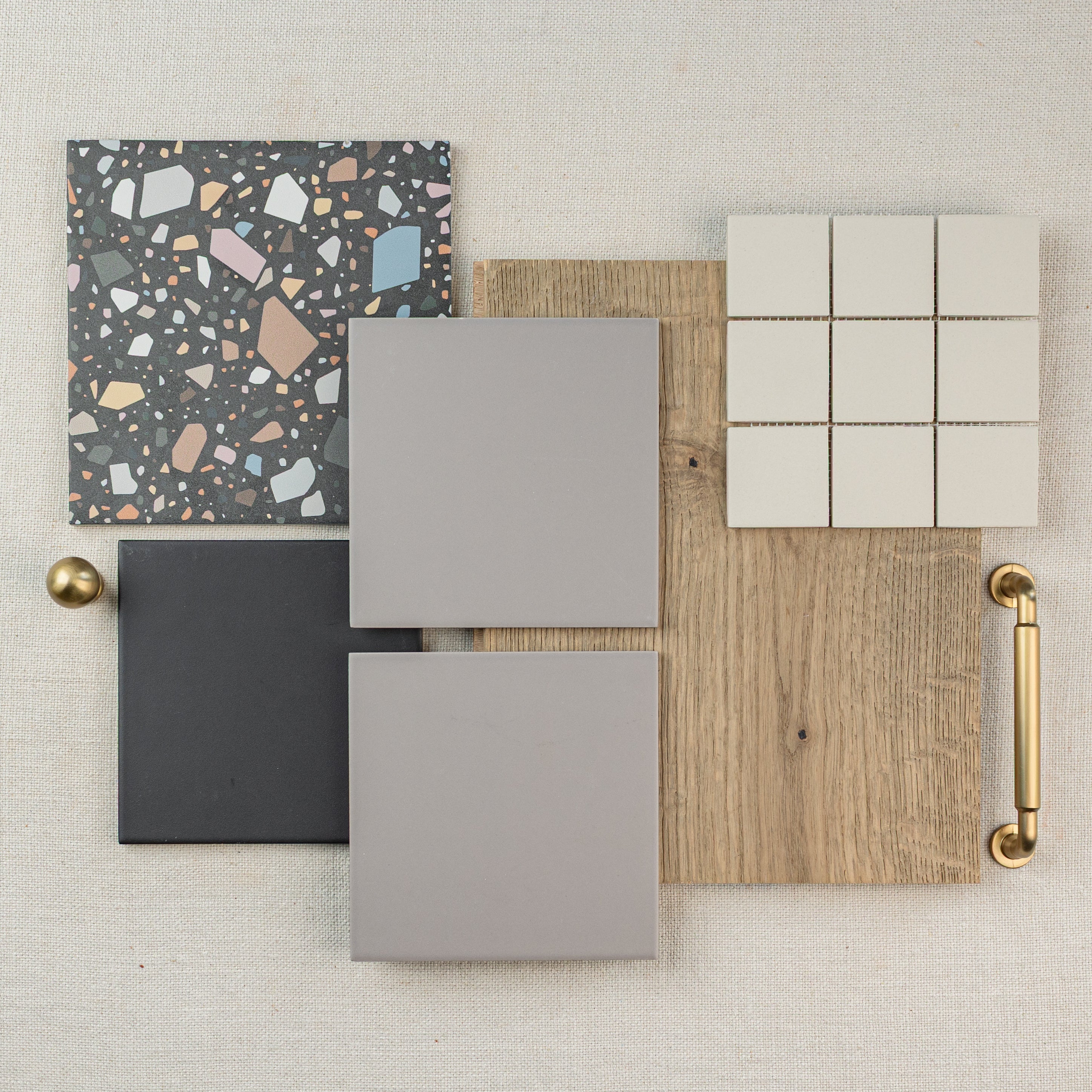 Flat lay of ceramic tiles in neutral tones, terrazzo tile, and a wood plank sample with a brass handle on fabric background
