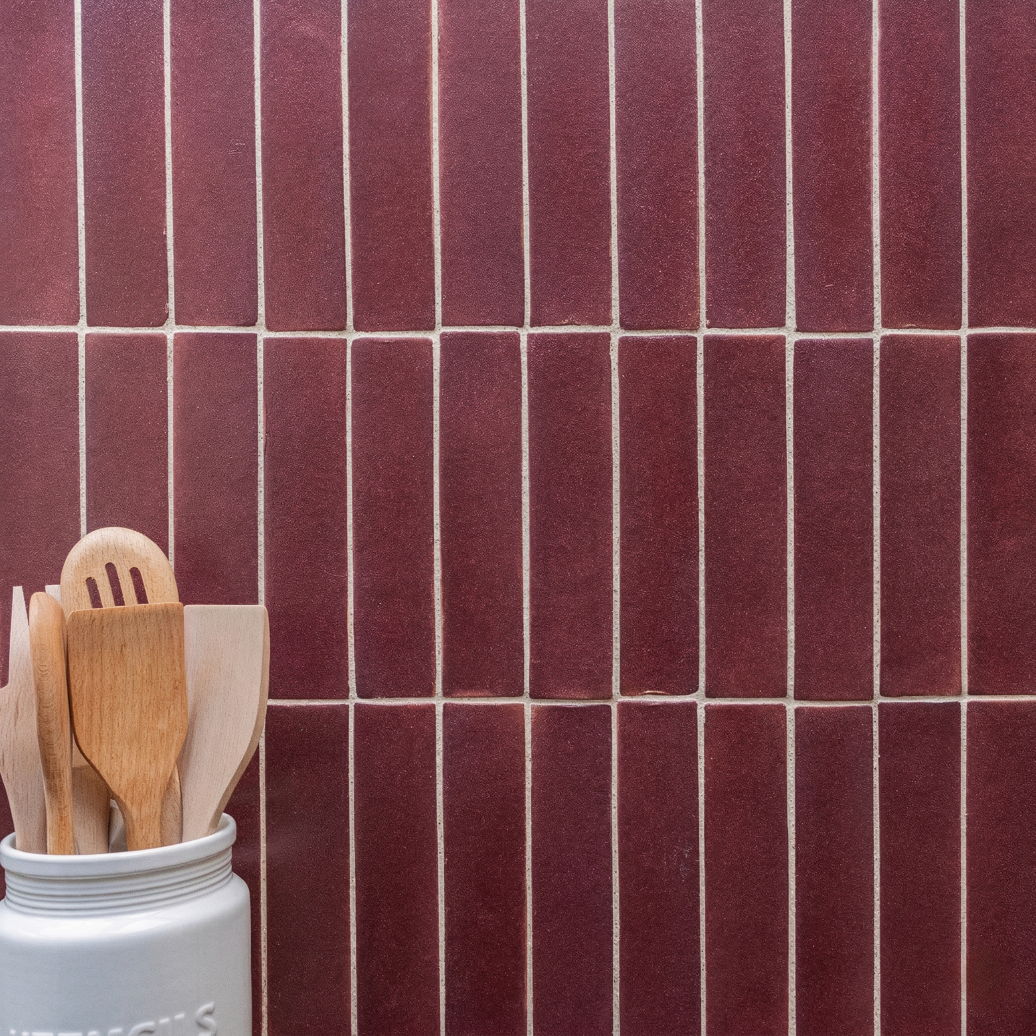 Vertical rectangular deep red ceramic tiles with white grout lines behind wooden kitchen utensils in a white jar