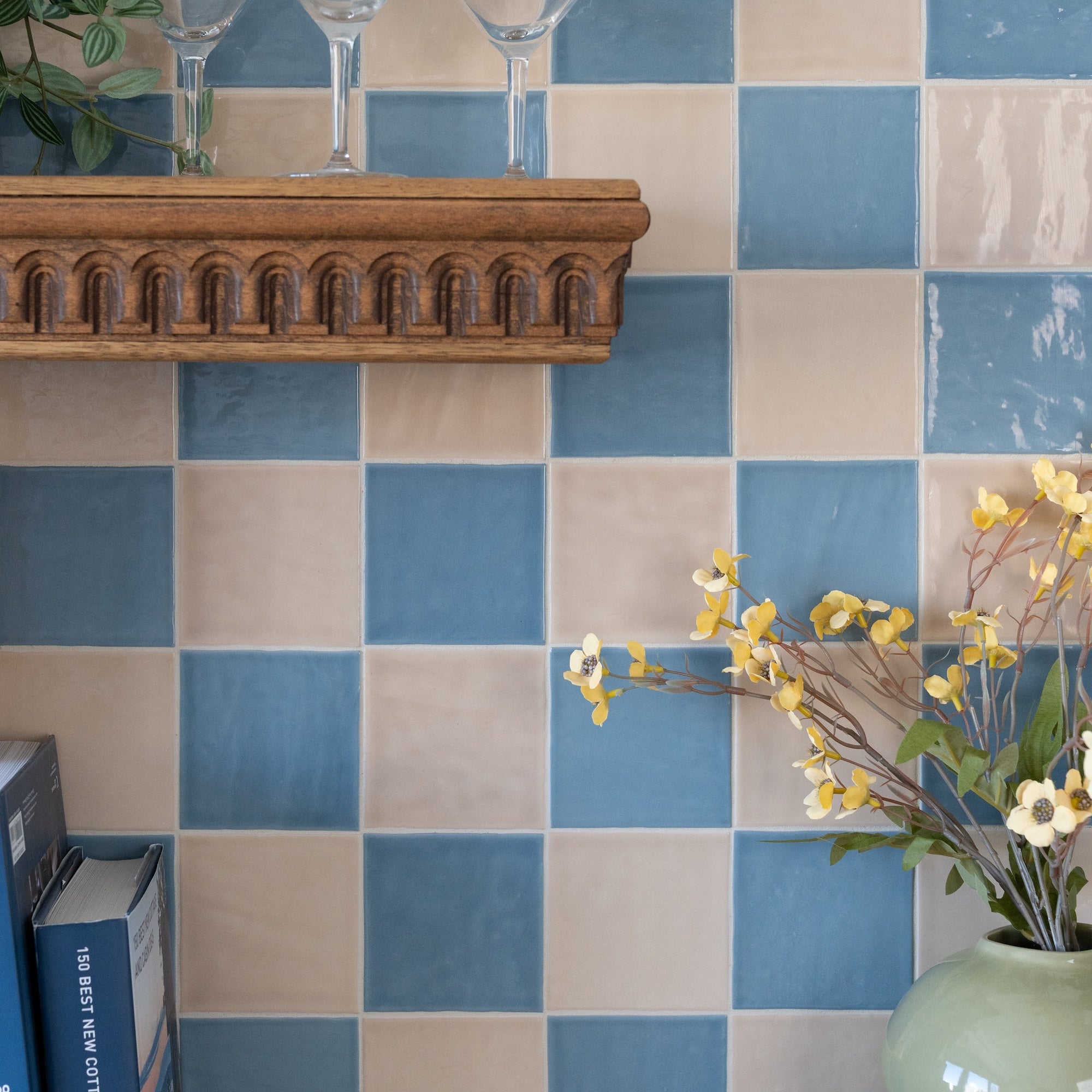 Blue and cream square tile wall with a carved wooden shelf, glasses, and flowers