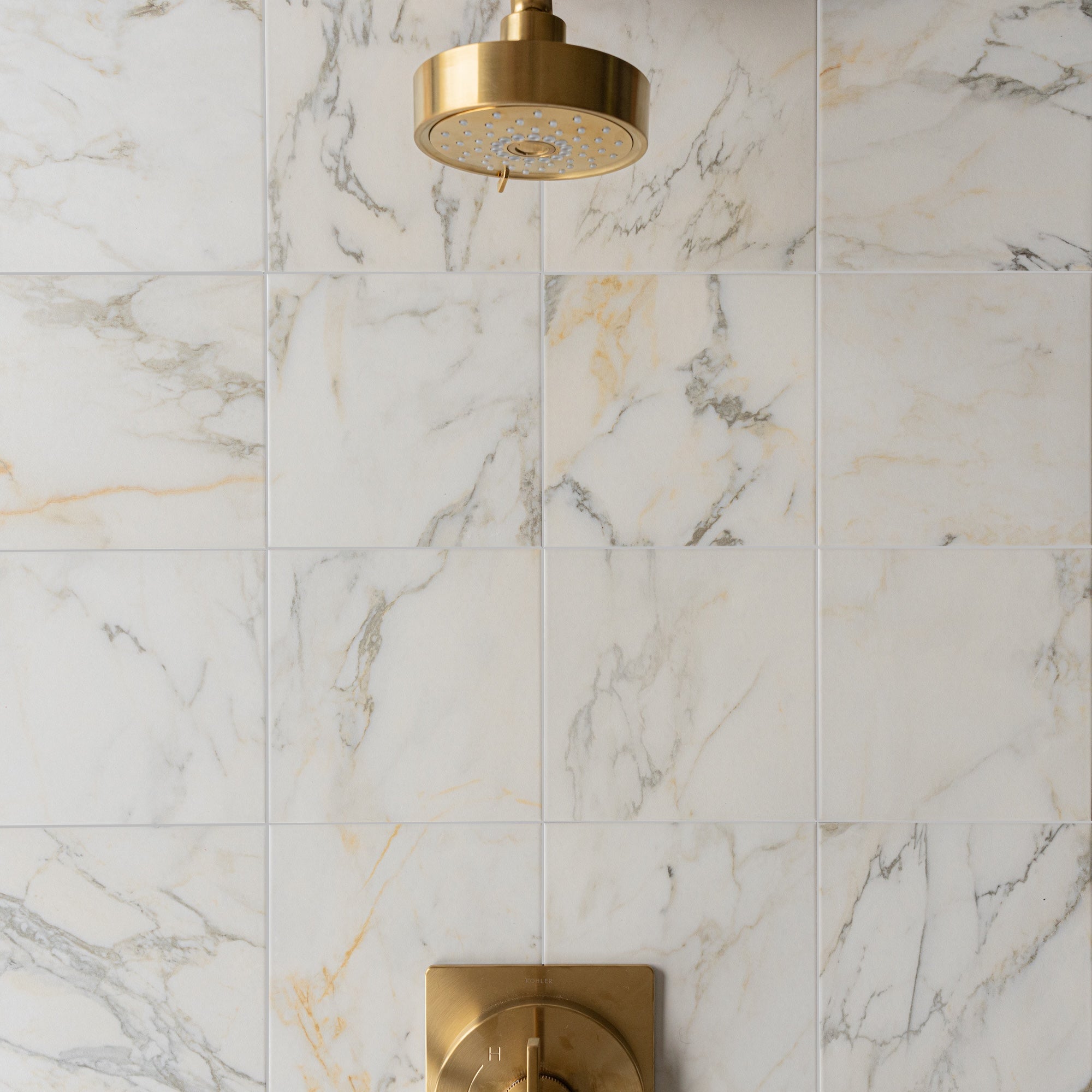 White and cream porcelain tiles with soft gray veining surrounding a modern gold showerhead and control knob