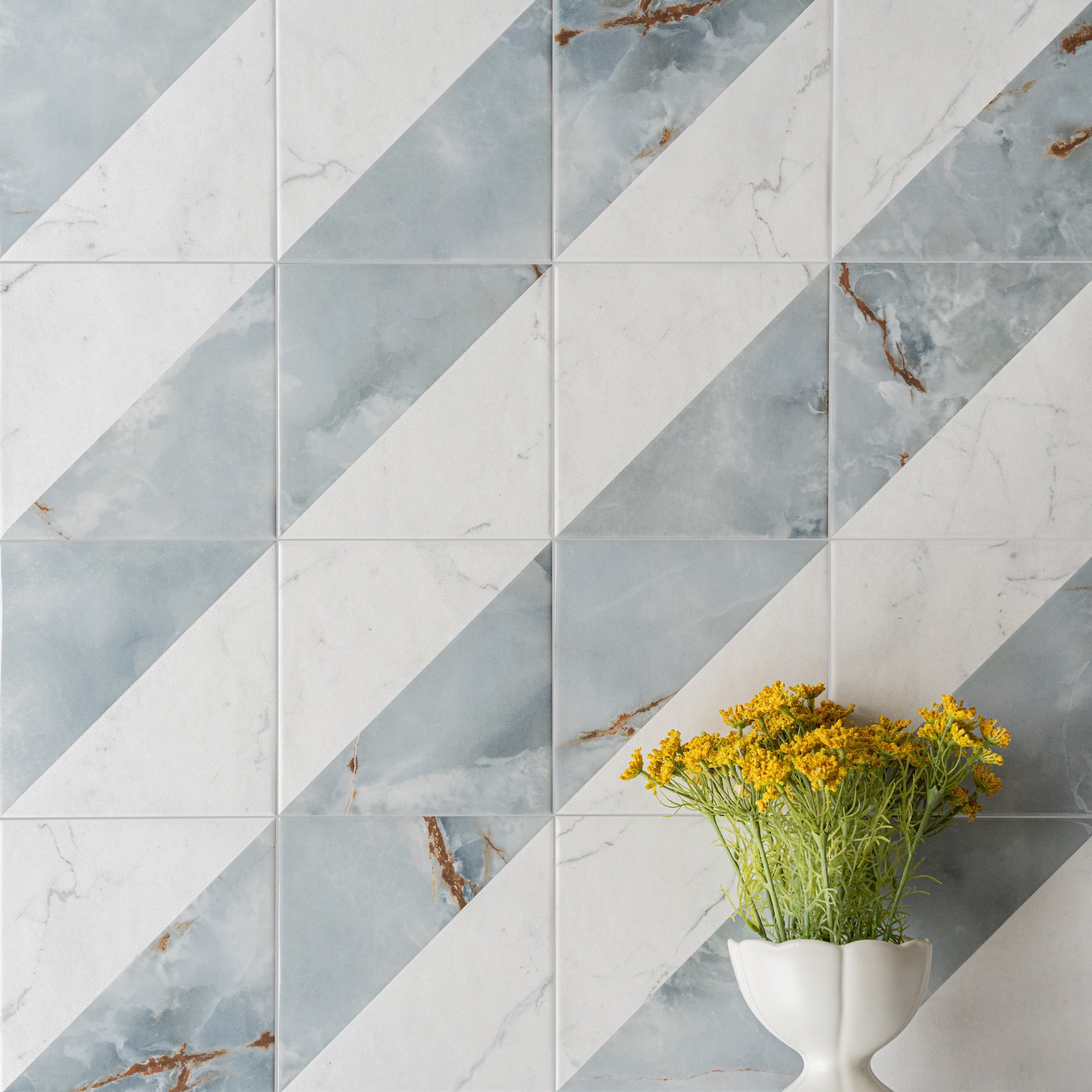 Blue and white marble-effect ceramic tiles arranged diagonally with a white vase holding yellow flowers in front