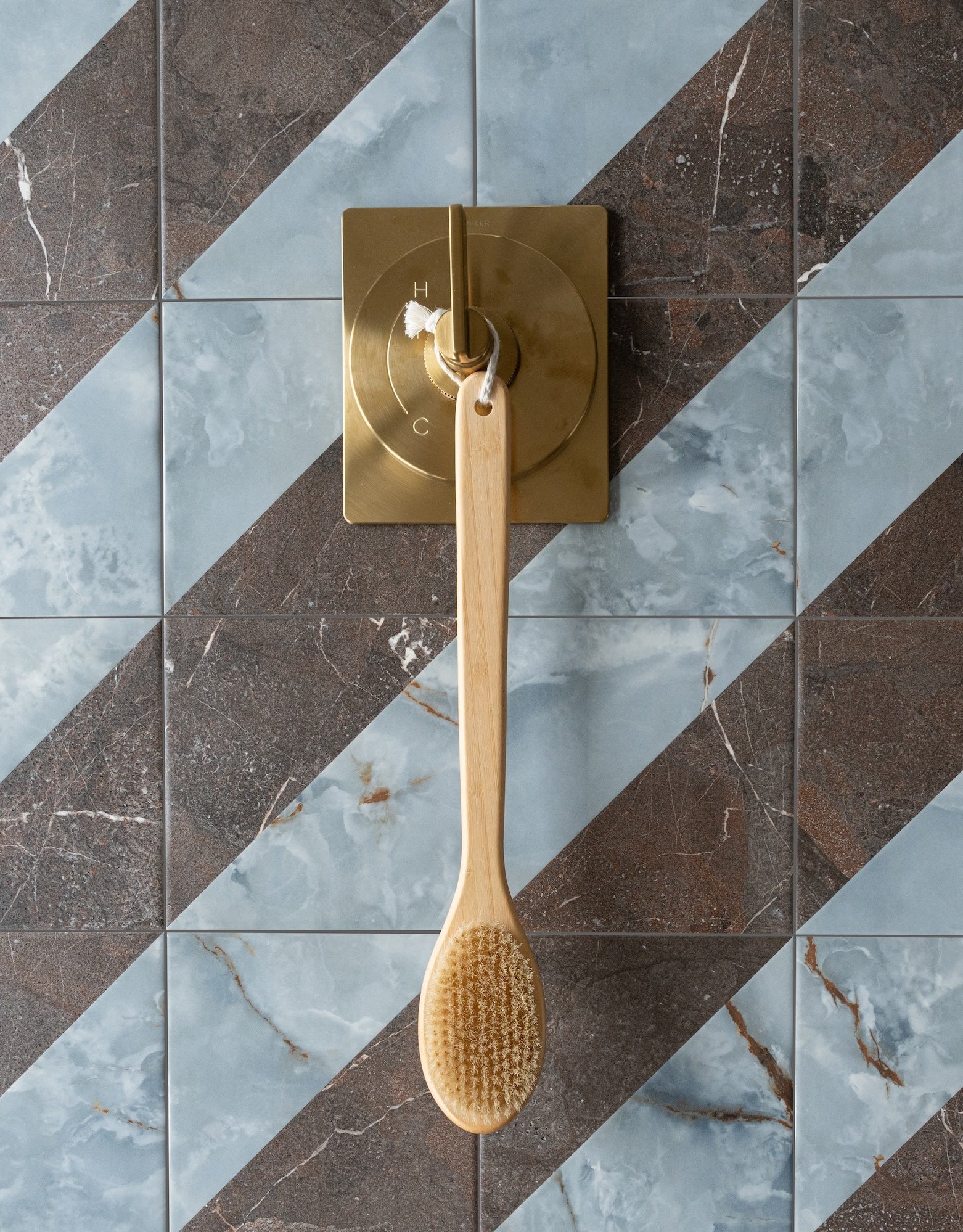 Long wooden bath brush hanging on a brass bathroom control against diagonally patterned blue and brown ceramic tiles