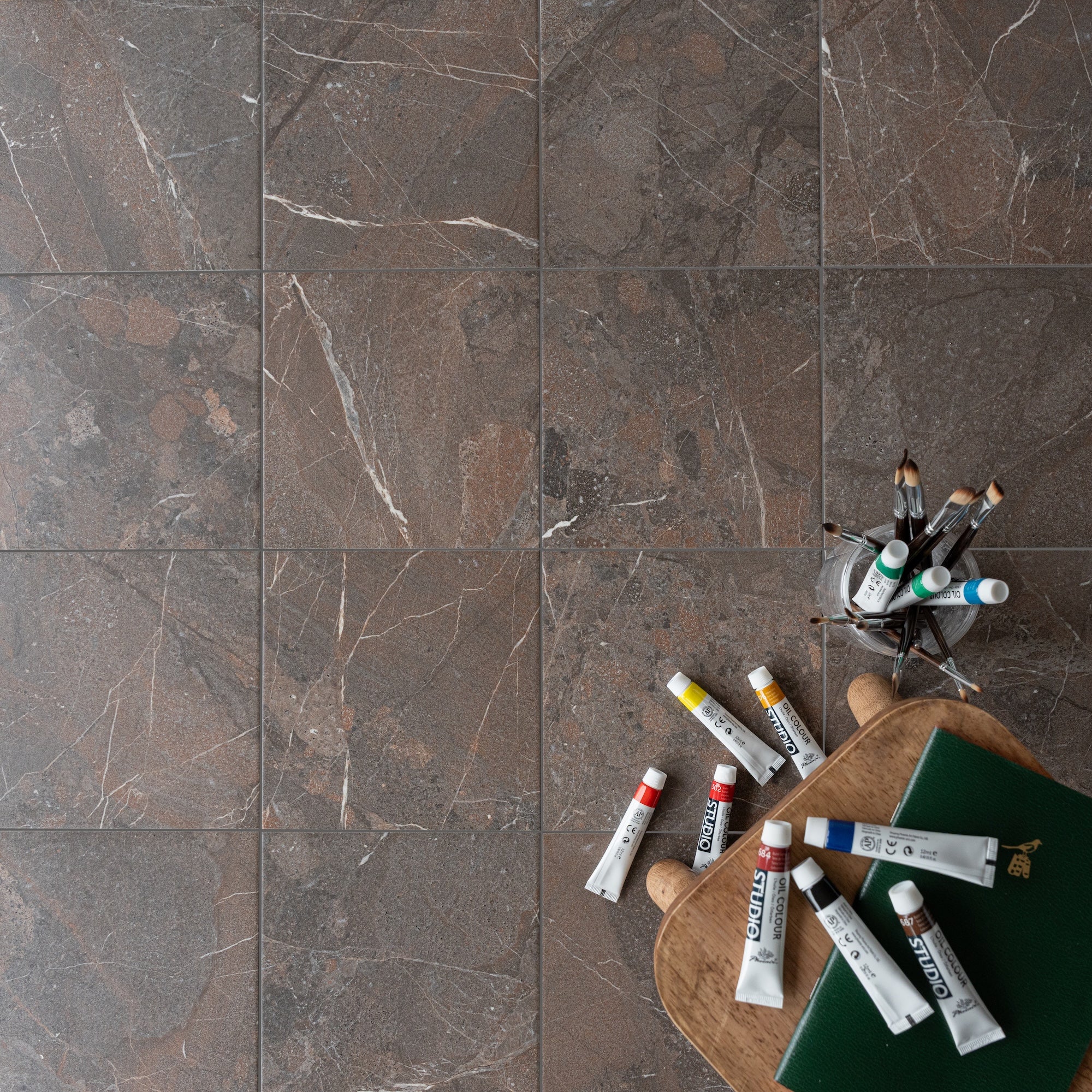 Overhead view of dark brown marble tiles with subtle white veining beside artist paint tubes and brushes on wooden board